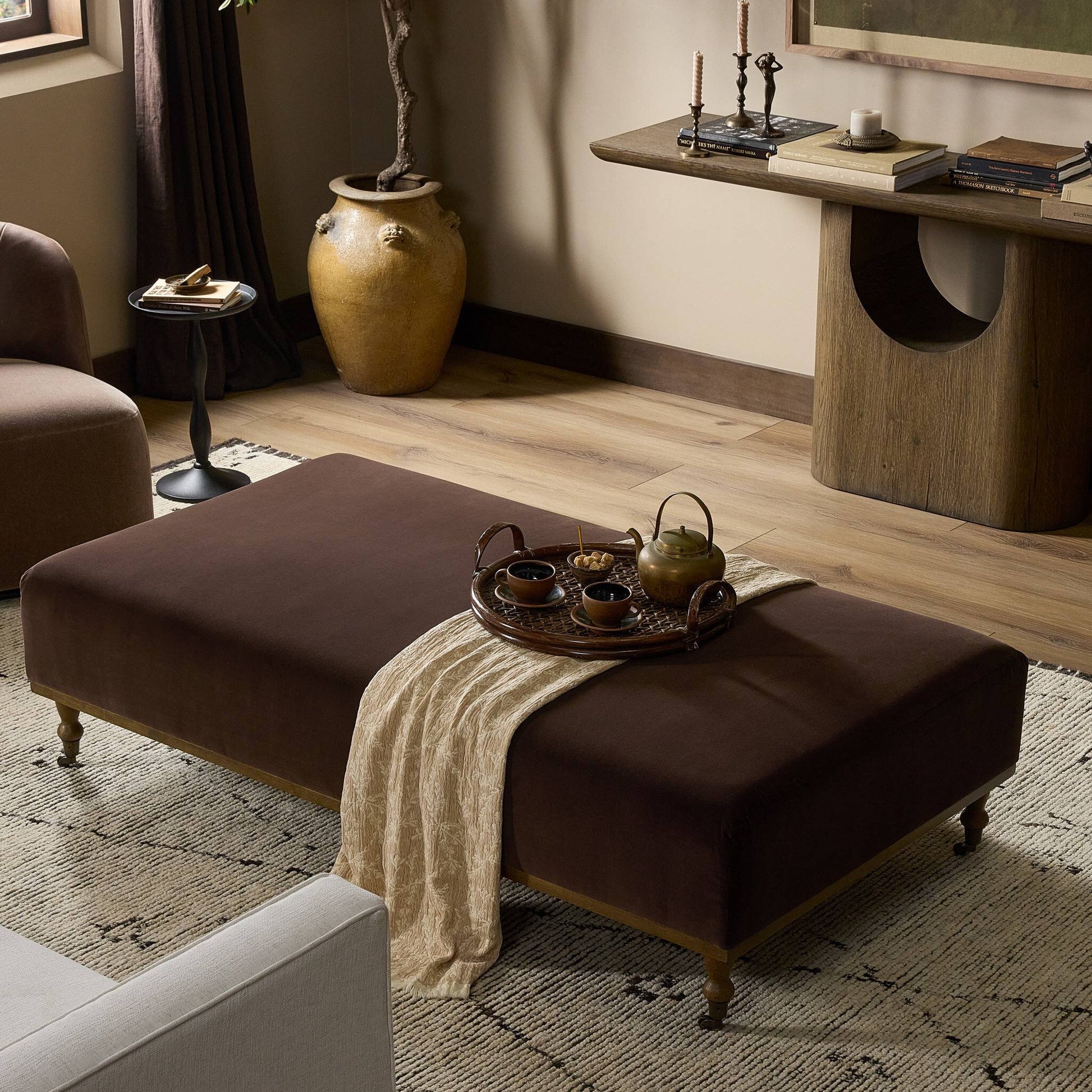 The Harlin Cocktail Ottoman, measuring 65.00"w x 35.00"d x 17.50"h with brown upholstery and wooden legs, serves as a coffee table topped with a tray holding a teapot and cups in a warmly decorated living room.