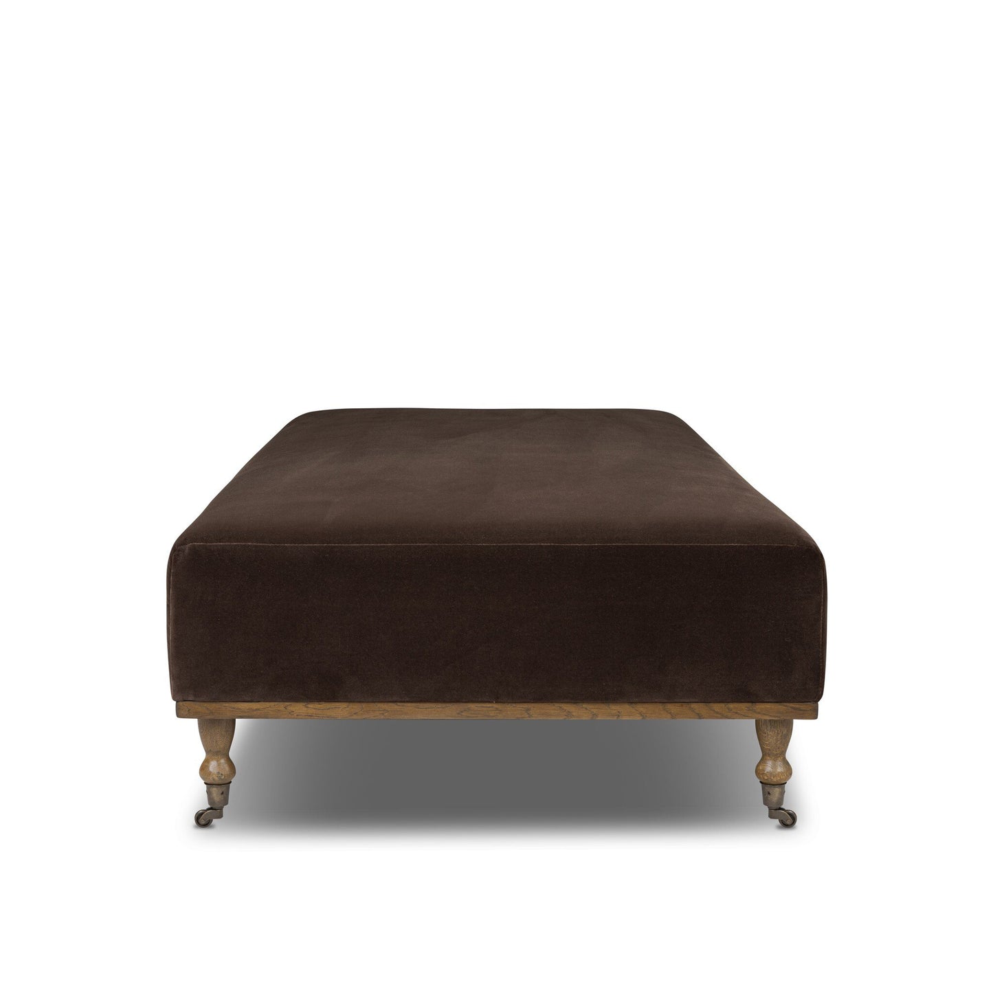 The Harlin Cocktail Ottoman features dark brown upholstery, wooden legs with front wheels, and measures 65"w x 35"d x 17.5"h. Shown against a white background.