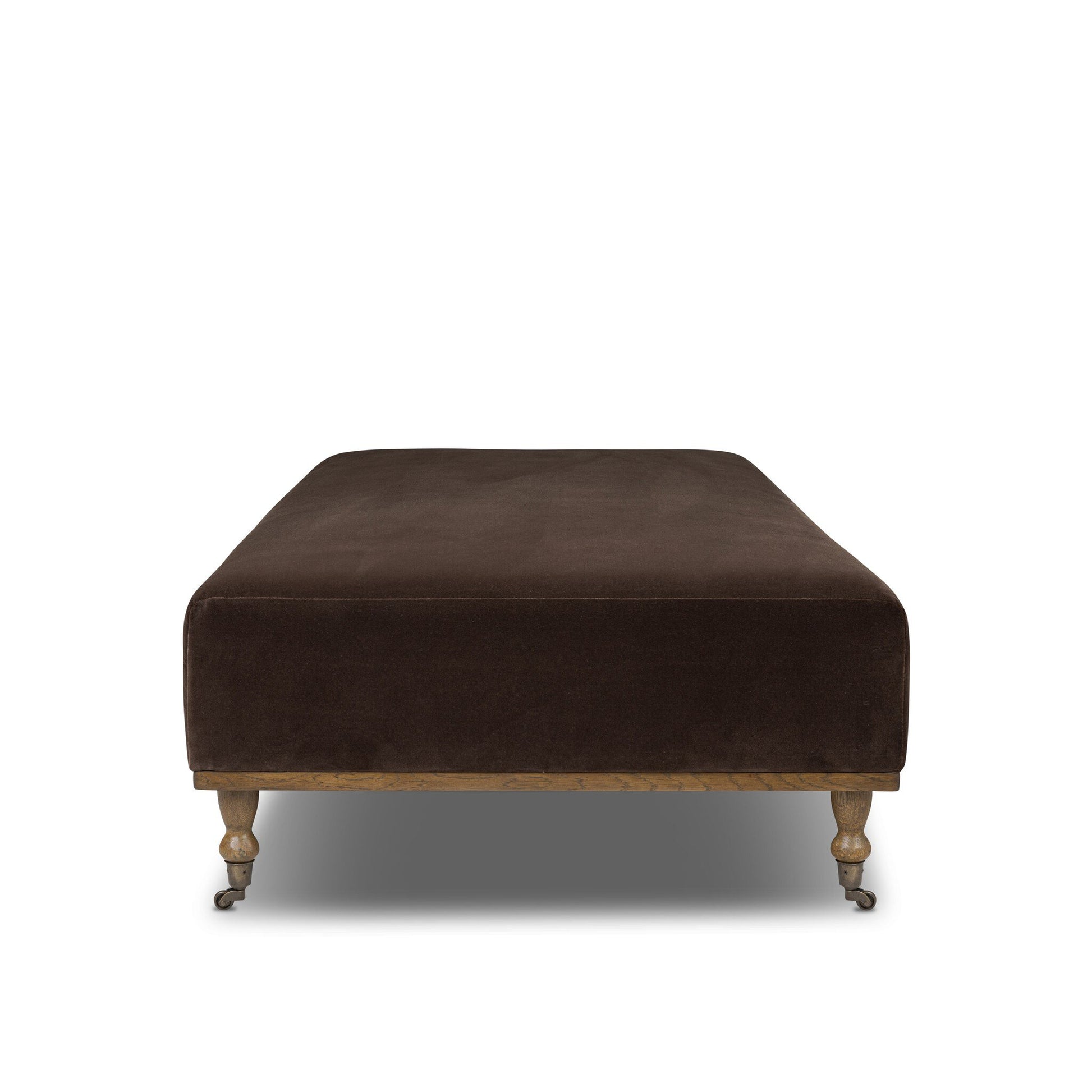 The Harlin Cocktail Ottoman features dark brown upholstery, wooden legs with front wheels, and measures 65"w x 35"d x 17.5"h. Shown against a white background.