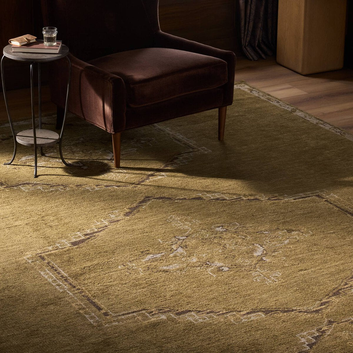 A brown upholstered armchair sits on an Astana Hand-Knotted 10'X14' rug next to a small round side table holding a glass of water and a book, in a warmly lit room with wooden floors and furniture.