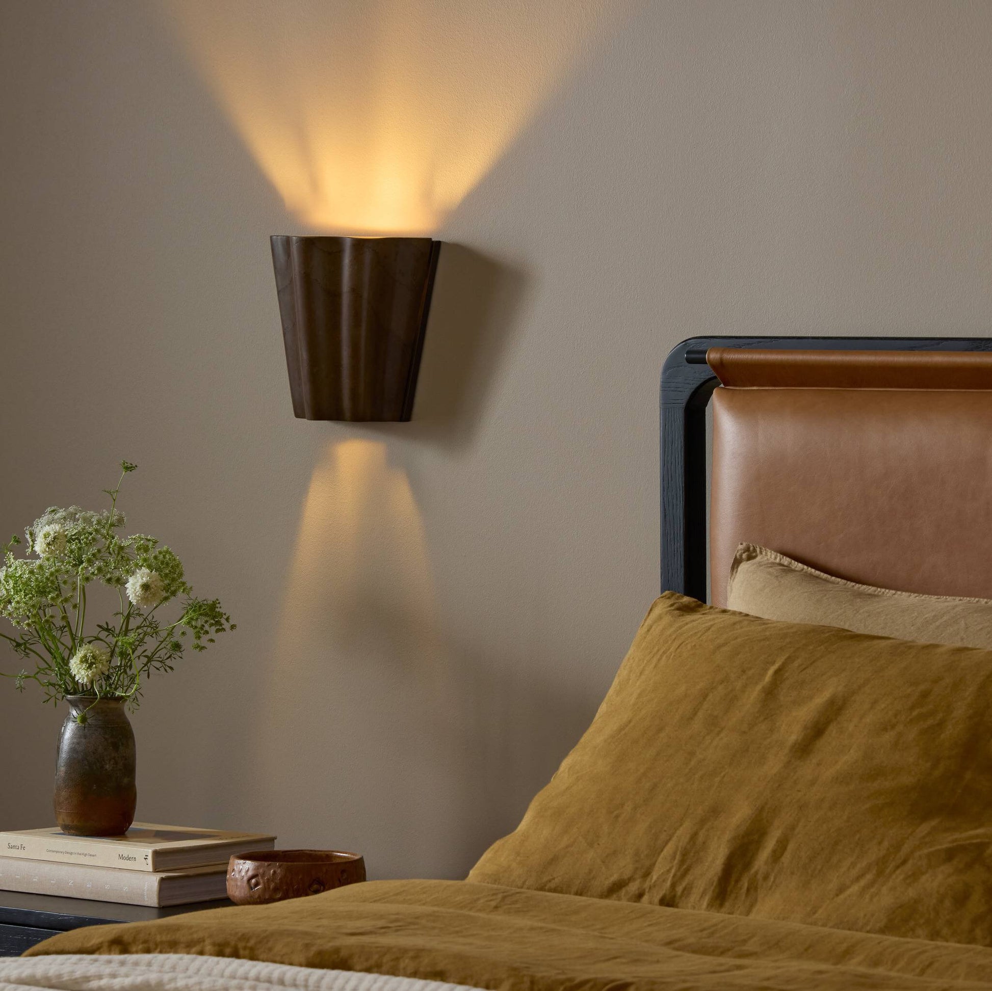 The Astra Sconce, measuring 8.50w x 4.50d x 7.75h in brown, casts warm light on a beige wall above a bed with brown and tan bedding. On the nightstand are a vase of flowers and several stacked books.