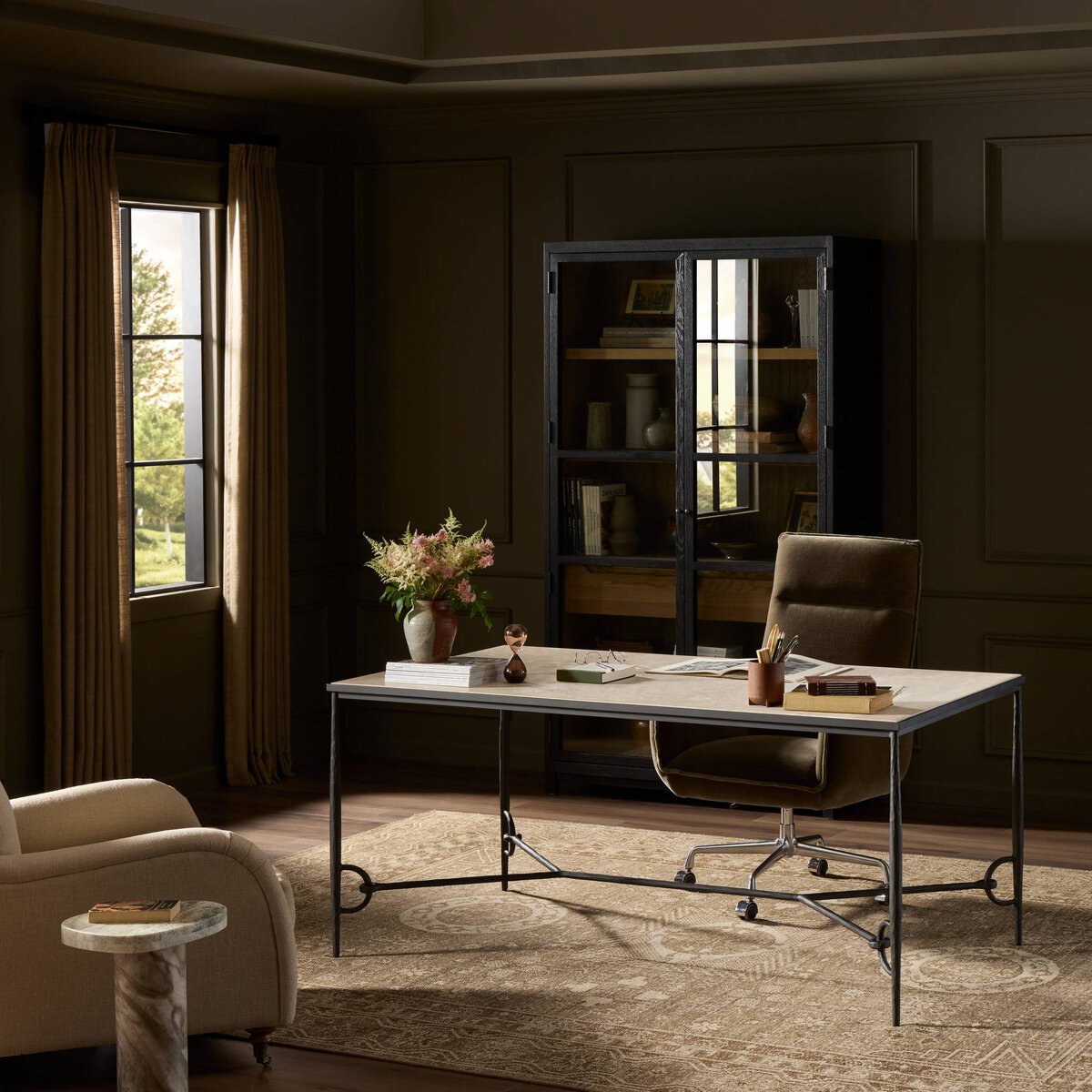 A stylish home office features the 68-inch wide Babylon Desk, a brown office chair, bookcase, beige armchair, marble side table, and tan curtains by the window; sunlight brightens the neutral walls and patterned rug.