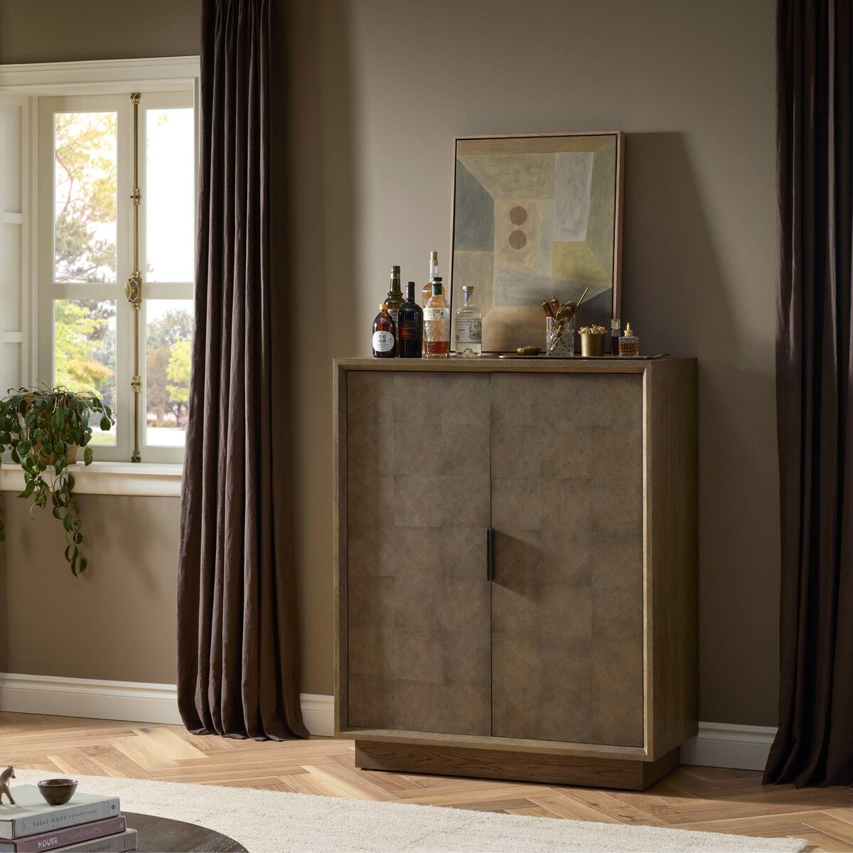 The Salta Bar Cabinet, featuring a modern geometric design, stands against a taupe wall and is topped with liquor bottles, glasses, and abstract art. Dimensions: 42.50"w x 20.00"d x 53.25"h. A window with drapes and a potted plant sit to the left.