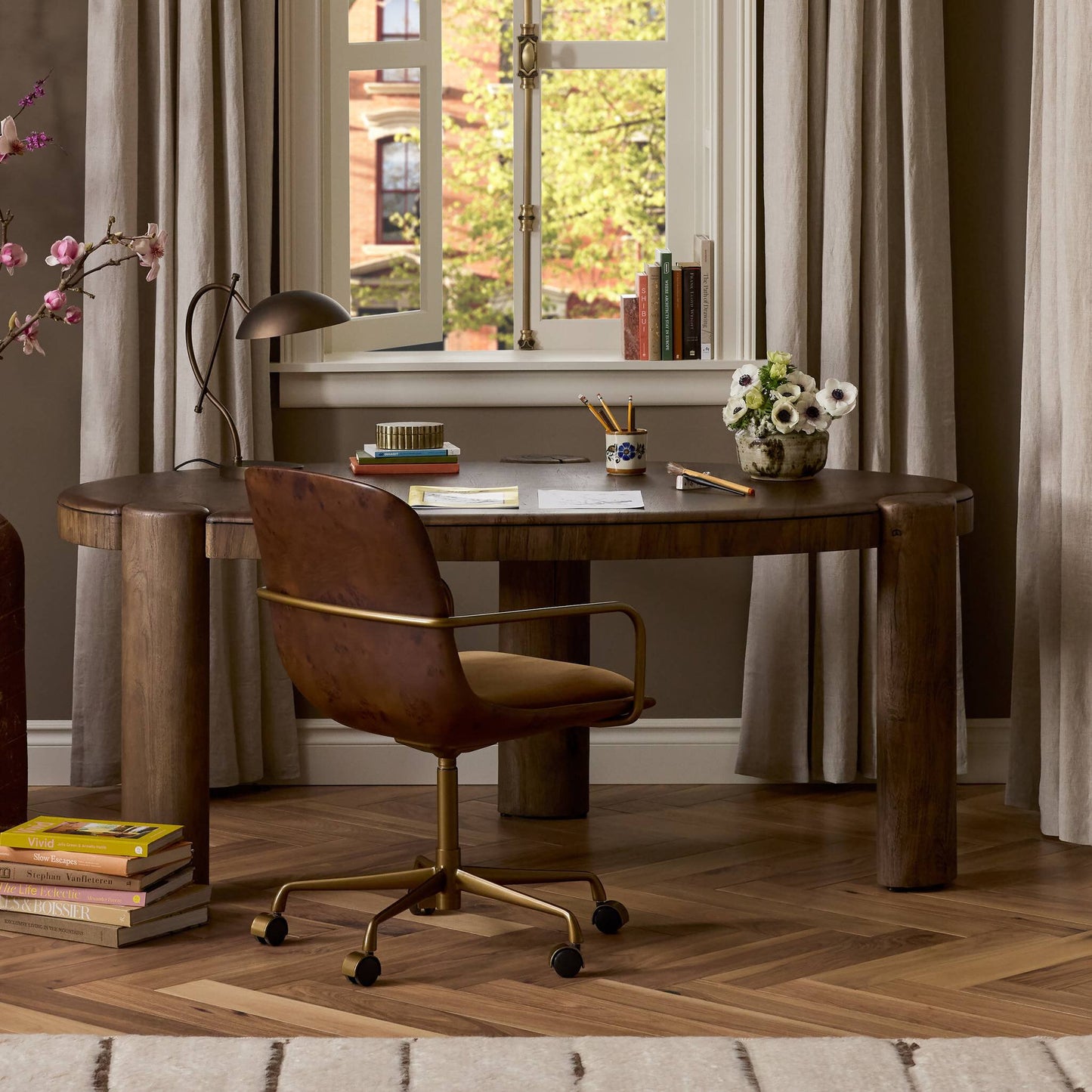 A stylish home office featuring the Kendra Desk Chair (21.50"w x 26.50"d x 36.25"h), a curved wooden desk, lamp, books, flowers, and papers by a window with beige curtains and a tree visible outside.