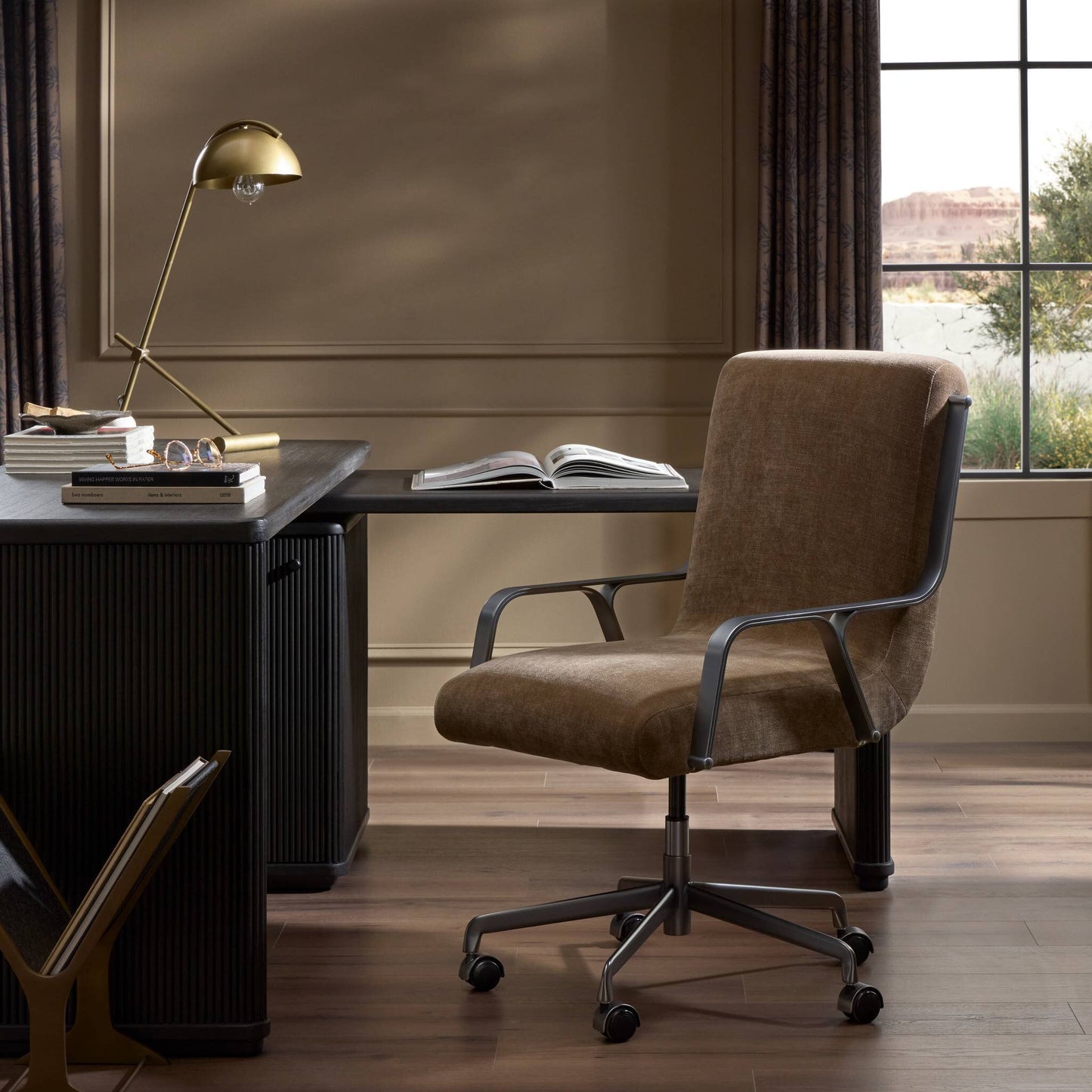 A modern home office featuring the beige upholstered Lou Desk Chair, a black desk, gold desk lamp, and an open book. A large window overlooks rocks and greenery. The room has wood floors and neutral walls.
