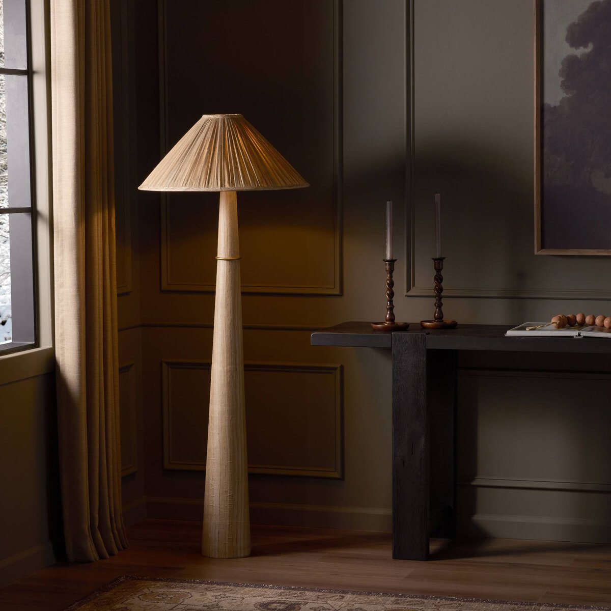 The Cordelia Floor Lamp, featuring a pleated shade and tall modern design, stands beside a dark wooden console table and office storage cabinet in an elegant paneled room, its light highlighting the decor through the nearby window.