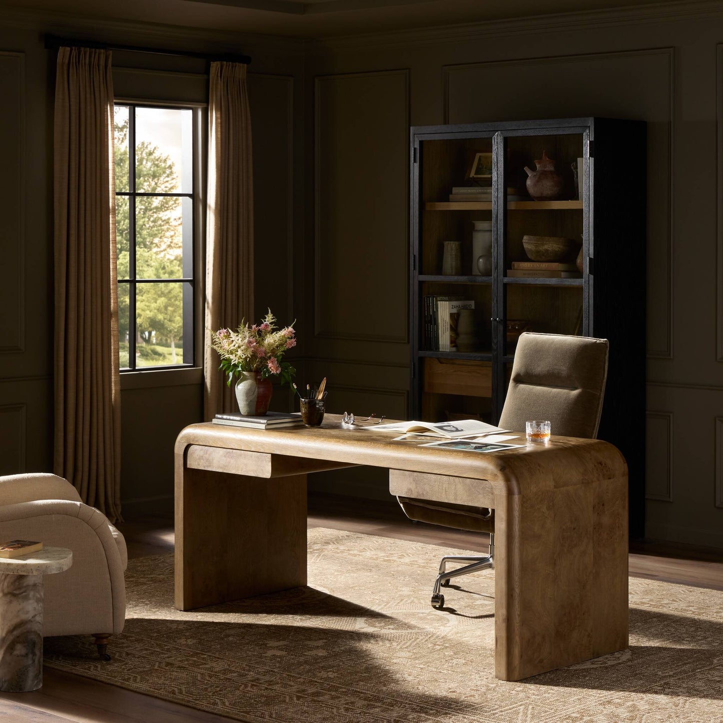 A cozy office with the modern Daven Desk (70"w x 28"d x 30"h), a swivel chair, bookshelf, and flower vase. Sunlight filters through beige curtains, highlighting the desk and neutral décor.