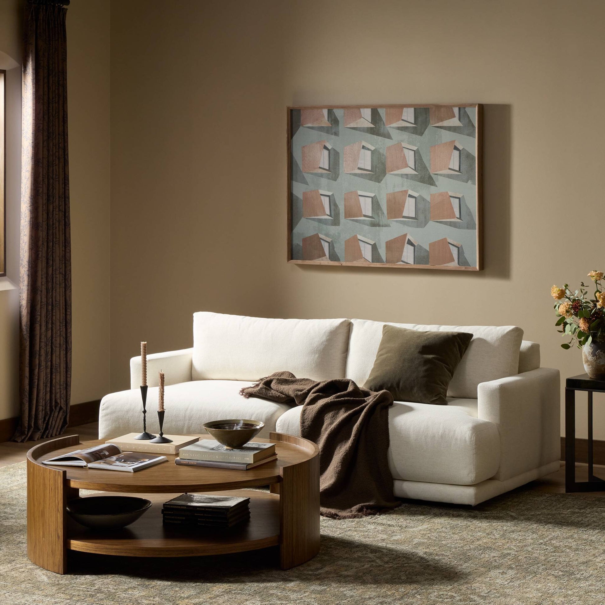 A modern living room with the 85" Jenning Sofa, styled with olive and brown cushions, a brown throw, a round wooden coffee table with decor, a vase of flowers, abstract art on the beige wall, and light streaming in from the left window.