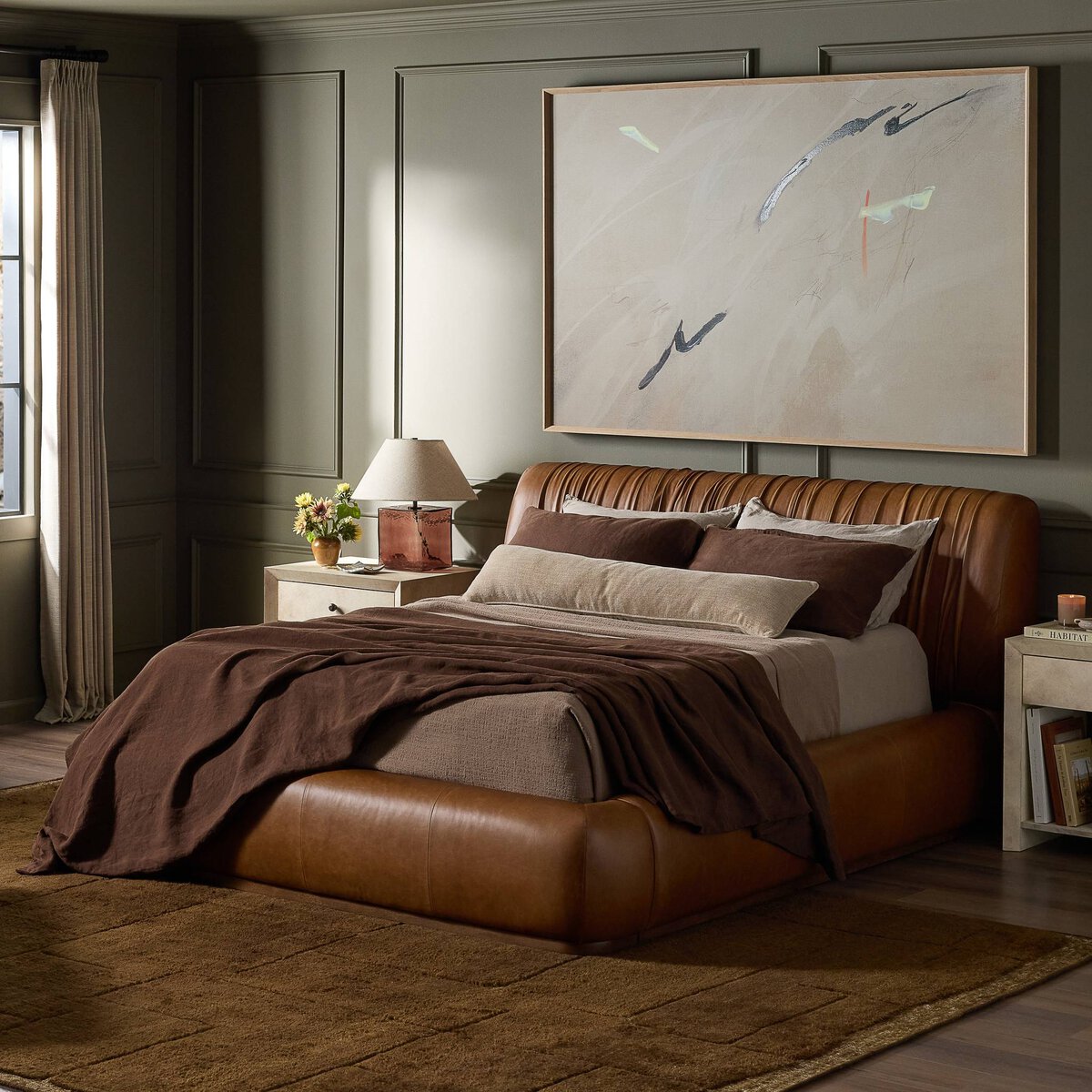 A modern bedroom featuring the Ronaldo Bed in brown leather (91.25"w x 101.00"d x 41.00"h), layered neutral bedding, two nightstands with lamps, a large abstract painting above the bed, and sunlight streaming through a window.