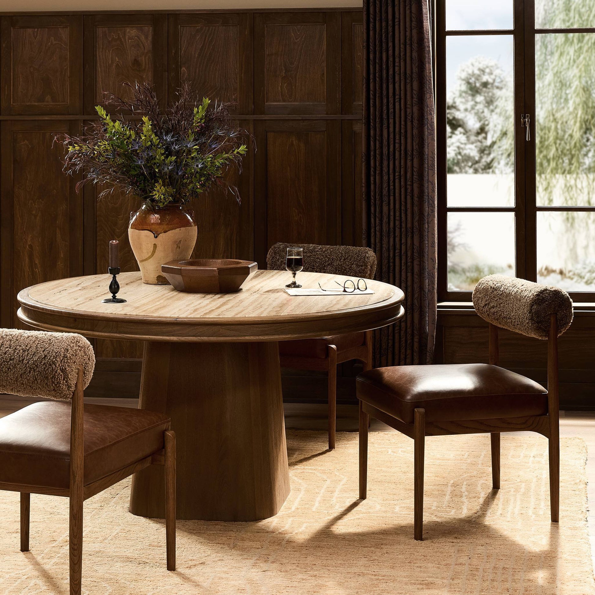 The Bourne Dining Table, measuring 55"x55"x30", displays a potted plant centerpiece. Four brown curved-back chairs rest on a beige rug, surrounded by wood paneling, dark curtains, and a large window.