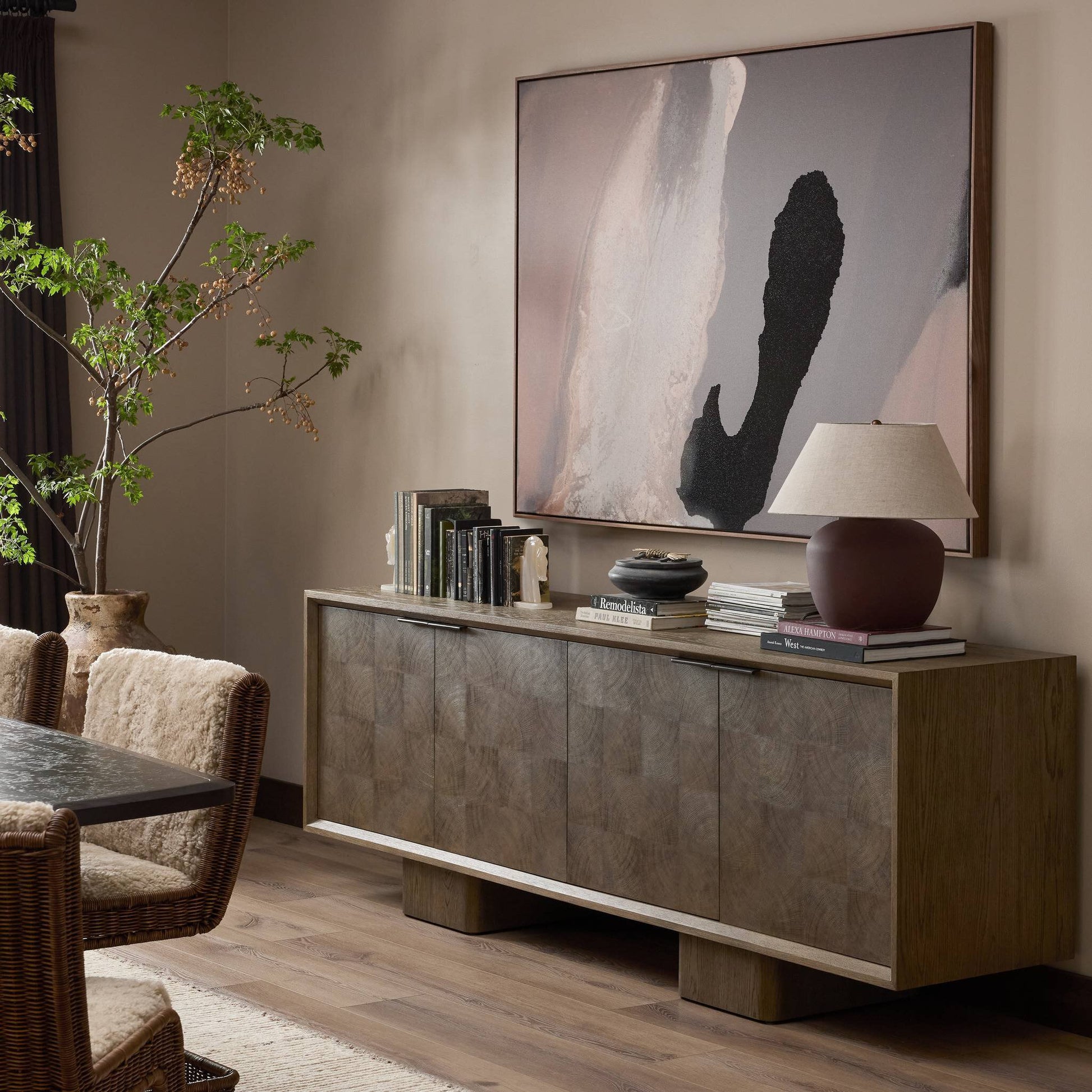 A modern living room features the Salta Sideboard (95.25"w x 20.00"d x 32.25"h) with books, a lamp, and decor, abstract art above, plus a dining table with textured chairs and a leafy plant on the wooden floor.