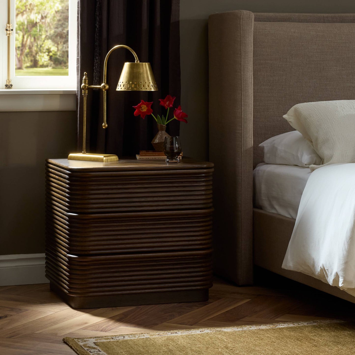 A modern bedroom features the Benny Nightstand (26.00"w x 22.00"d x 24.00"h) in brown wood, topped with a brass lamp, red flowers, water glass, and books next to a beige upholstered bed by a sunlit window.