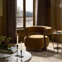 The Yuri Swivel Chair, a cozy and modern mustard-yellow seat (34.25"w x 31.25"d x 28.25"h), sits by a window with long curtains in a warmly lit room, next to a round side table holding a drink and book, with candles and flowers on the coffee table nearby.