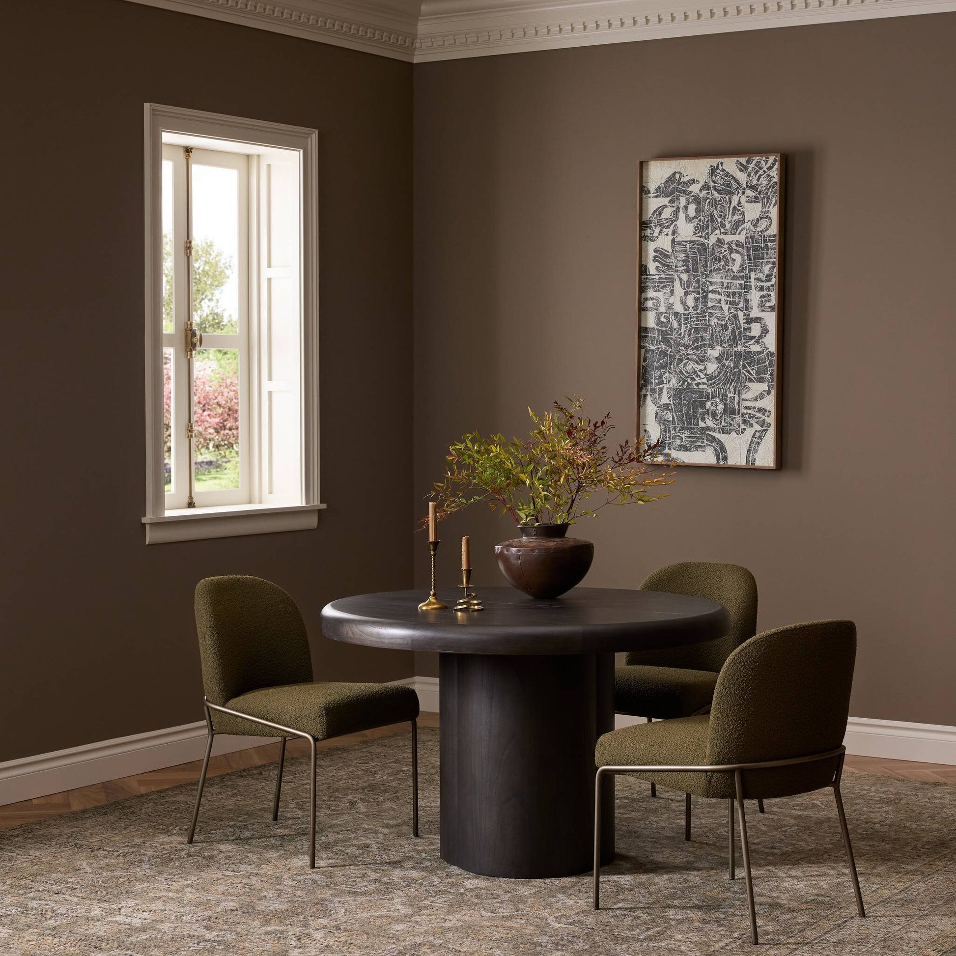 The Pemble Extension Dining Table (48x48) with two green cushioned chairs sits in a brown-walled room, decorated with a vase of branches, candlesticks, framed art, and bright natural light streaming through the window.