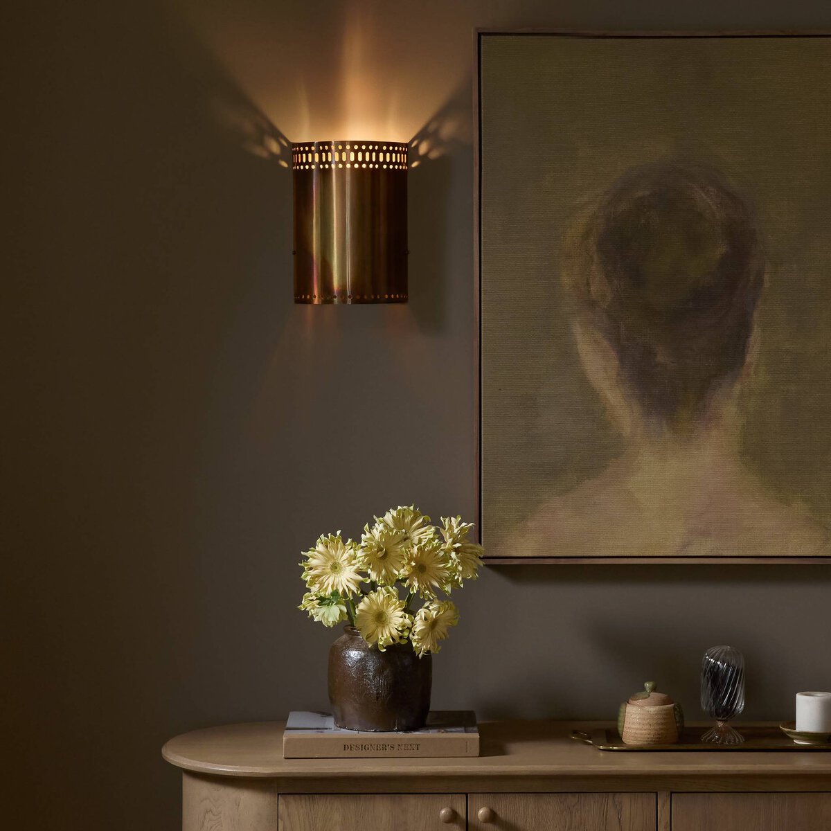 A softly lit room features the Rialto Sconce in brass, a painting of a person's head from behind, a wooden sideboard, a brown vase with yellow flowers, acid wash accents, and warm minimalist decor.