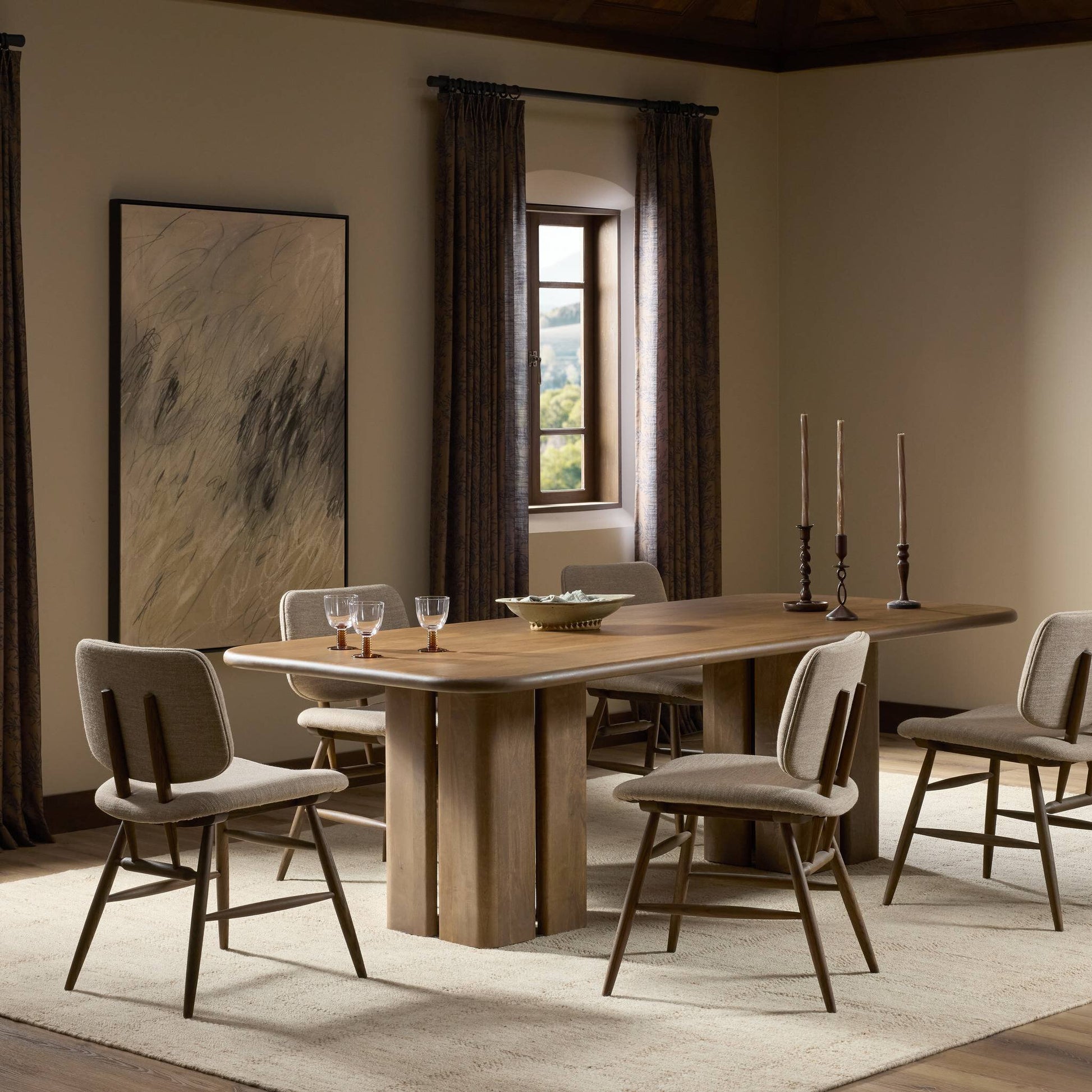 A modern dining room showcases the Rishi Dining Table with six upholstered chairs, three tall candlesticks, a bowl, and glasses. Beige walls, a large abstract painting, and dark curtained windows create an elegant atmosphere.