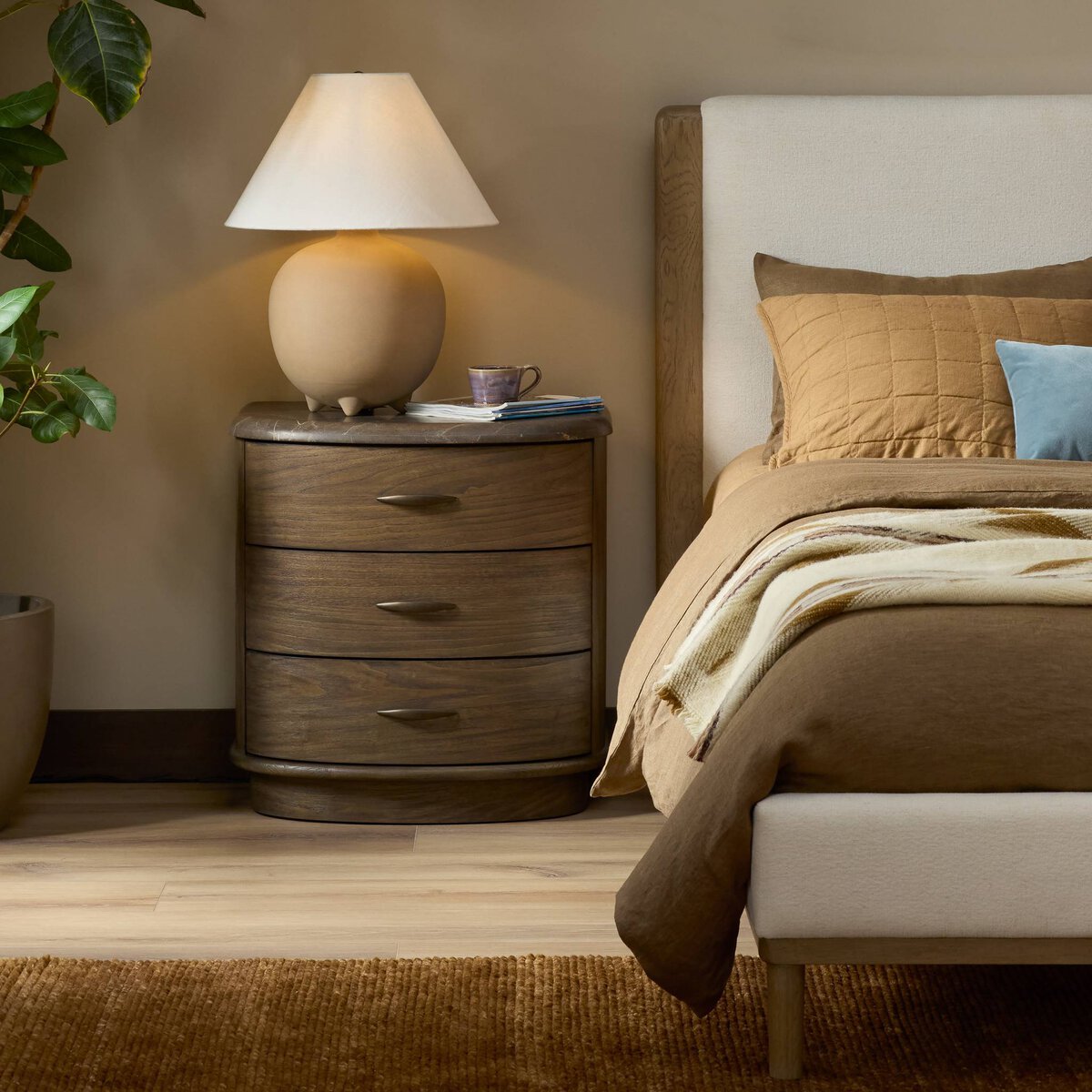A cozy bedroom features a Tovae Nightstand (26.00"w x 18.50"d x 26.50"h) with a beige lamp, purple cup, and book. The bed has tan and brown bedding, a light blue accent pillow, and a green plant on the left side.
