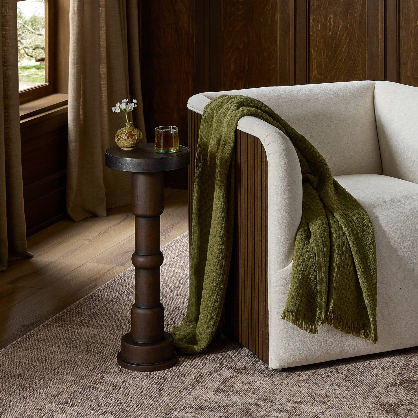 A cozy cream armchair with a green textured throw sits beside the Mizu End Table (10x10x24.75), which holds a vase and drink in a warmly lit room with wood-paneled walls and a large window.