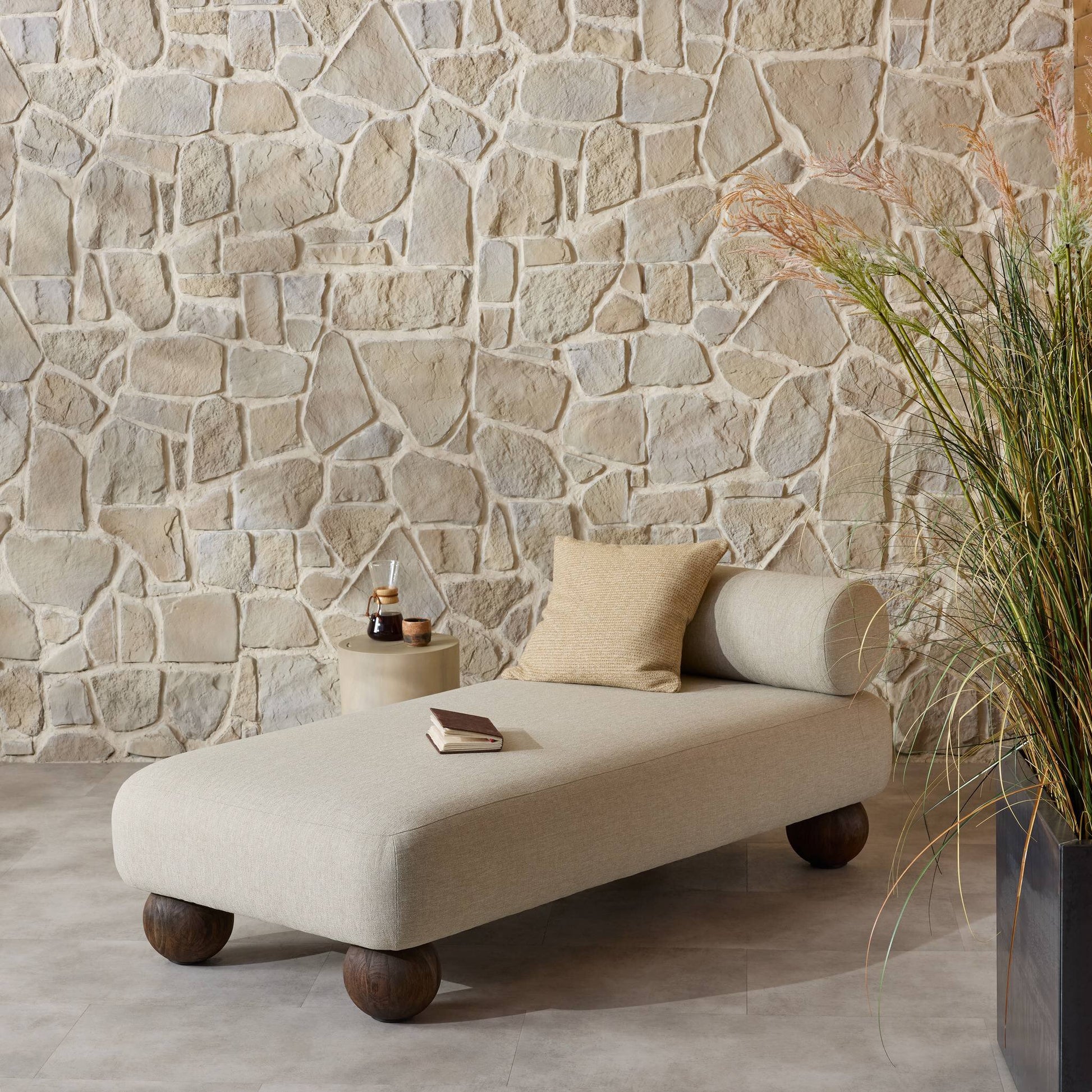 The Leilani Outdoor Chaise with a pillow and book sits on a tiled floor by a stone wall, next to a round side table with a drink and book. An elegant office desk is nearby, with tall ornamental grass positioned to the right.