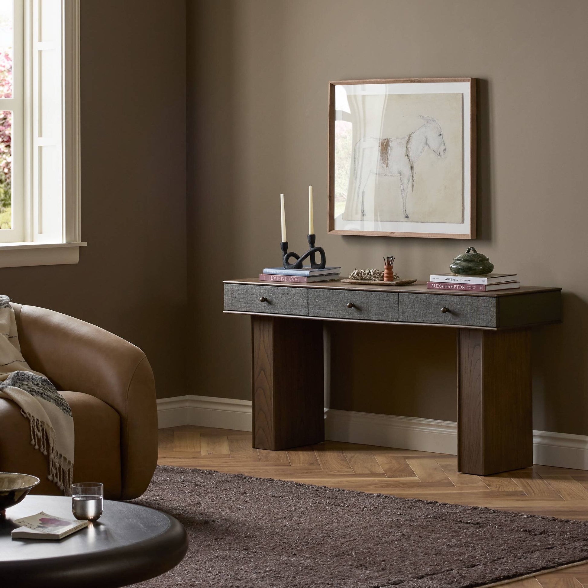 A minimalist living room corner with a brown sofa and the Soloman Console Table, which has three drawers, books, two candles, a small sculpture, and a framed horse sketch displayed on the taupe wall above.
