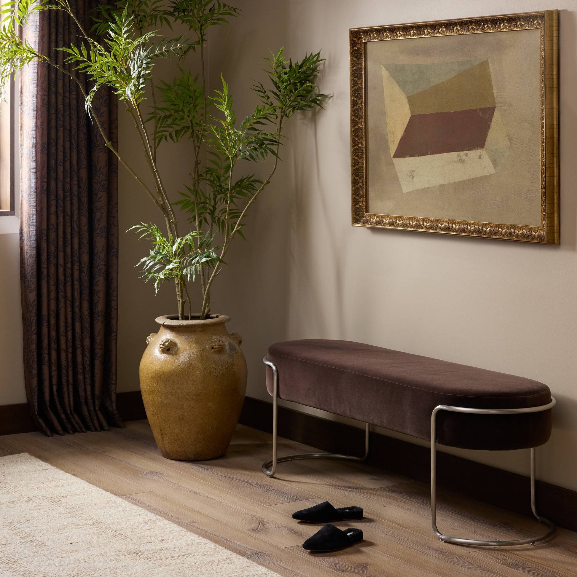 A stylish room corner features the 52.50"w Savoy Accent Bench with metal legs, a tall potted plant in a large brown vase, black slippers on the floor, and a geometric painting on the wall. Natural light streams through a curtained window.