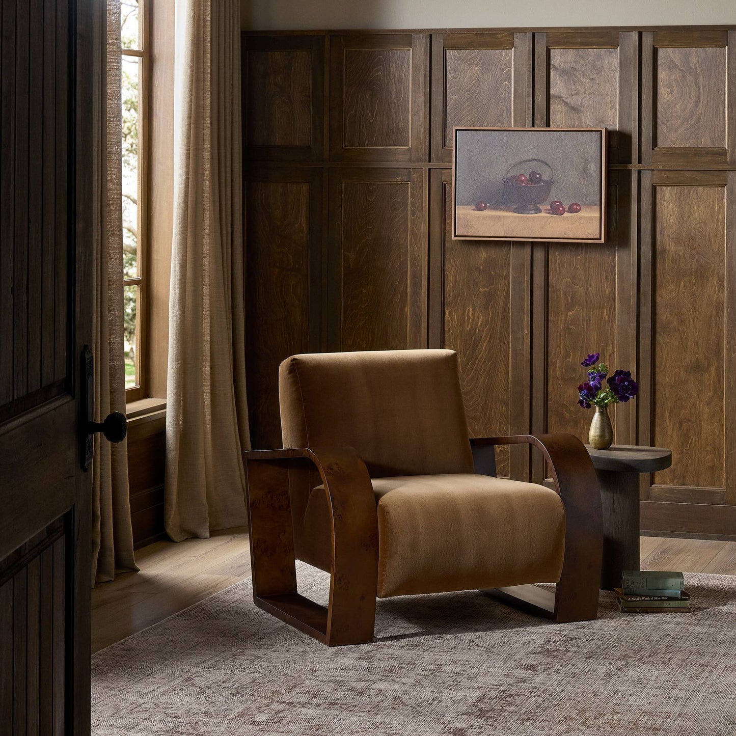 A cozy room with wood-paneled walls, a large window with beige curtains, the comfortable Montez Chair upholstered in brown, a small round table with a vase of purple flowers, and a framed still life painting on the wall.