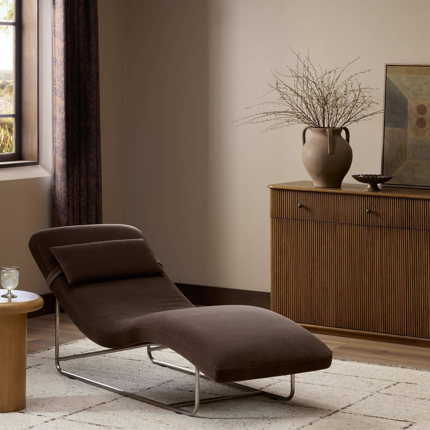 A modern living room featuring the dark brown Michi Chaise (27x65"), a round wood side table with a glass of water, wooden cabinet with vase and branches, abstract art, and sunlight streaming through curtained windows.