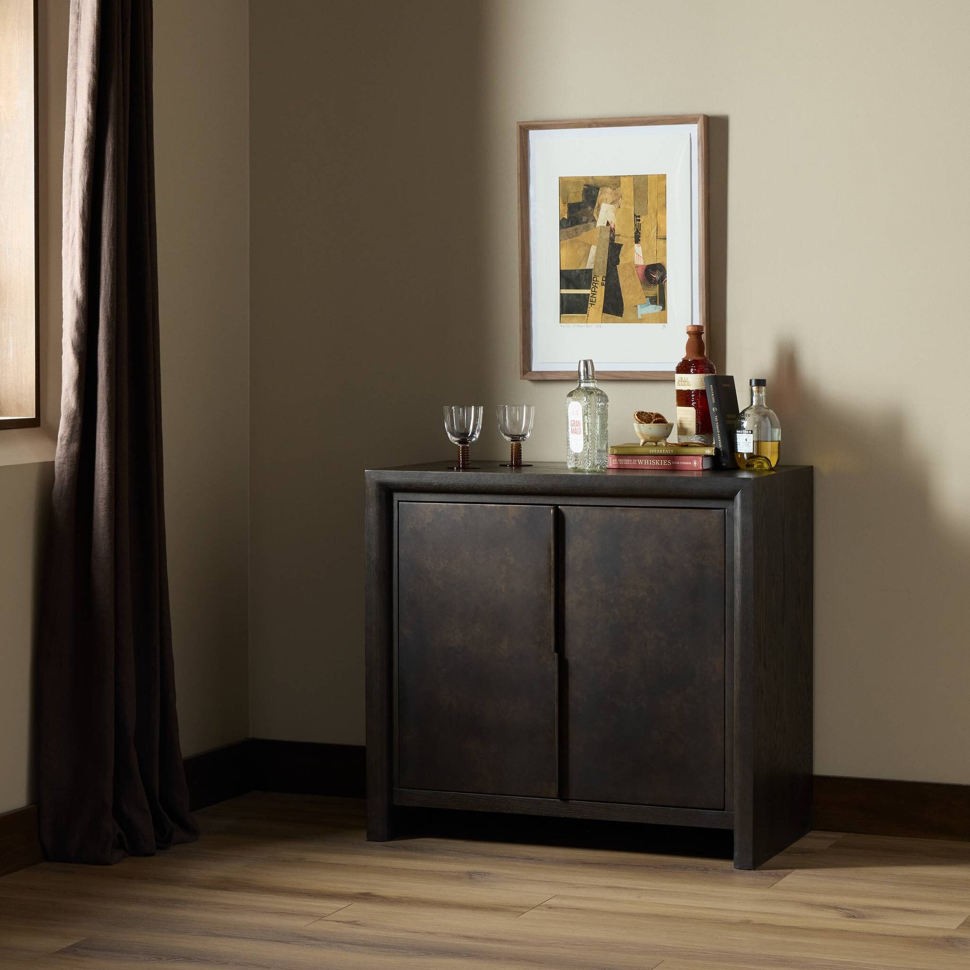 The Arlington Small Cabinet, made of dark wood, stores glasses and liquor bottles. A small bowl and decor sit on top. An abstract artwork hangs above; sunlight streams through a window with brown curtains onto wood floors and light beige walls.