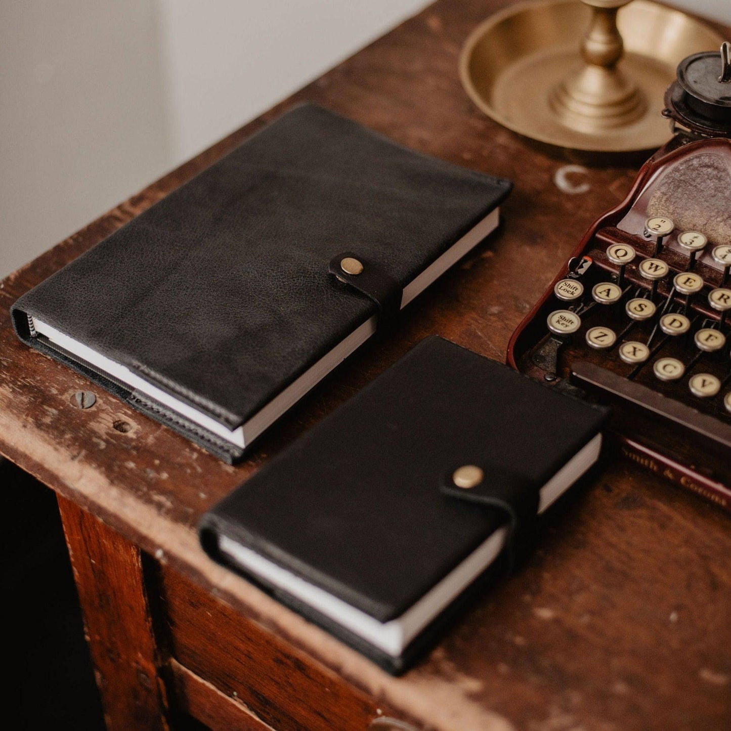 Two black Writer's Journals with button closures rest on a wooden desk beside a vintage typewriter and brass dish, each handcrafted as a custom branded keepsake.