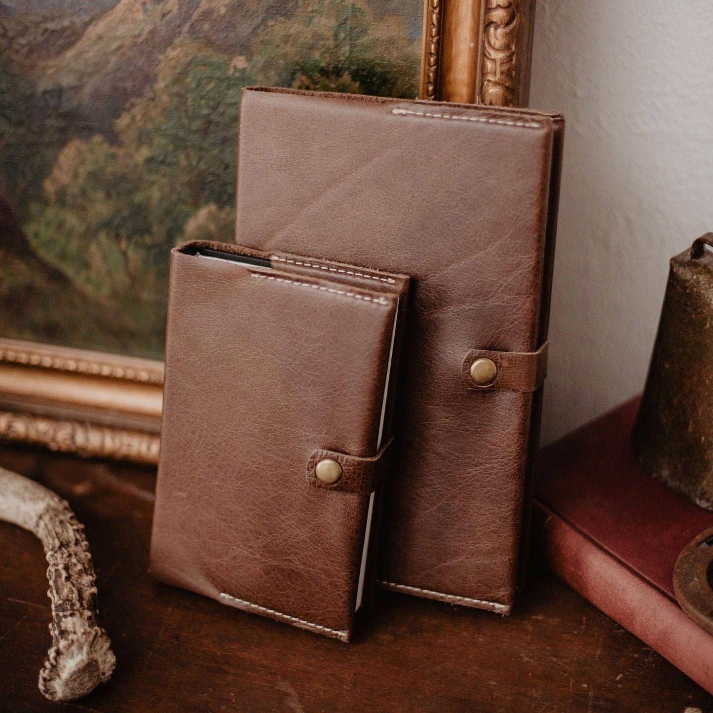 Two handcrafted Writer's Journals with button clasps, one larger than the other, stand upright on a wooden surface in front of a framed painting and vintage decor—timeless keepsakes made in the USA, ready to be cherished.