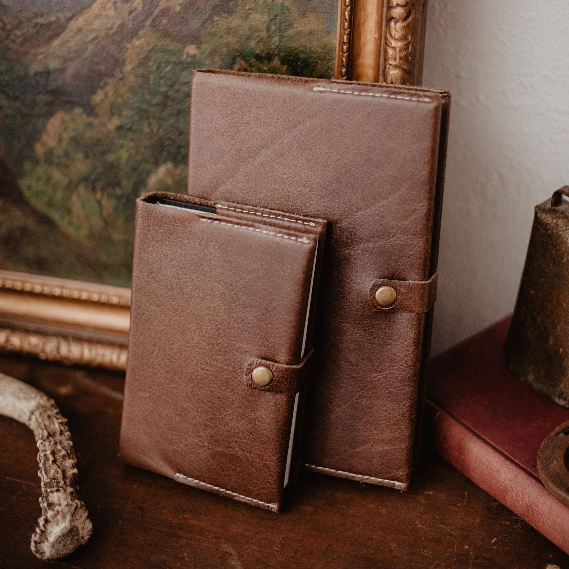 Two handcrafted Writer's Journals with button clasps, one larger than the other, stand upright on a wooden surface in front of a framed painting and vintage decor—timeless keepsakes made in the USA, ready to be cherished.