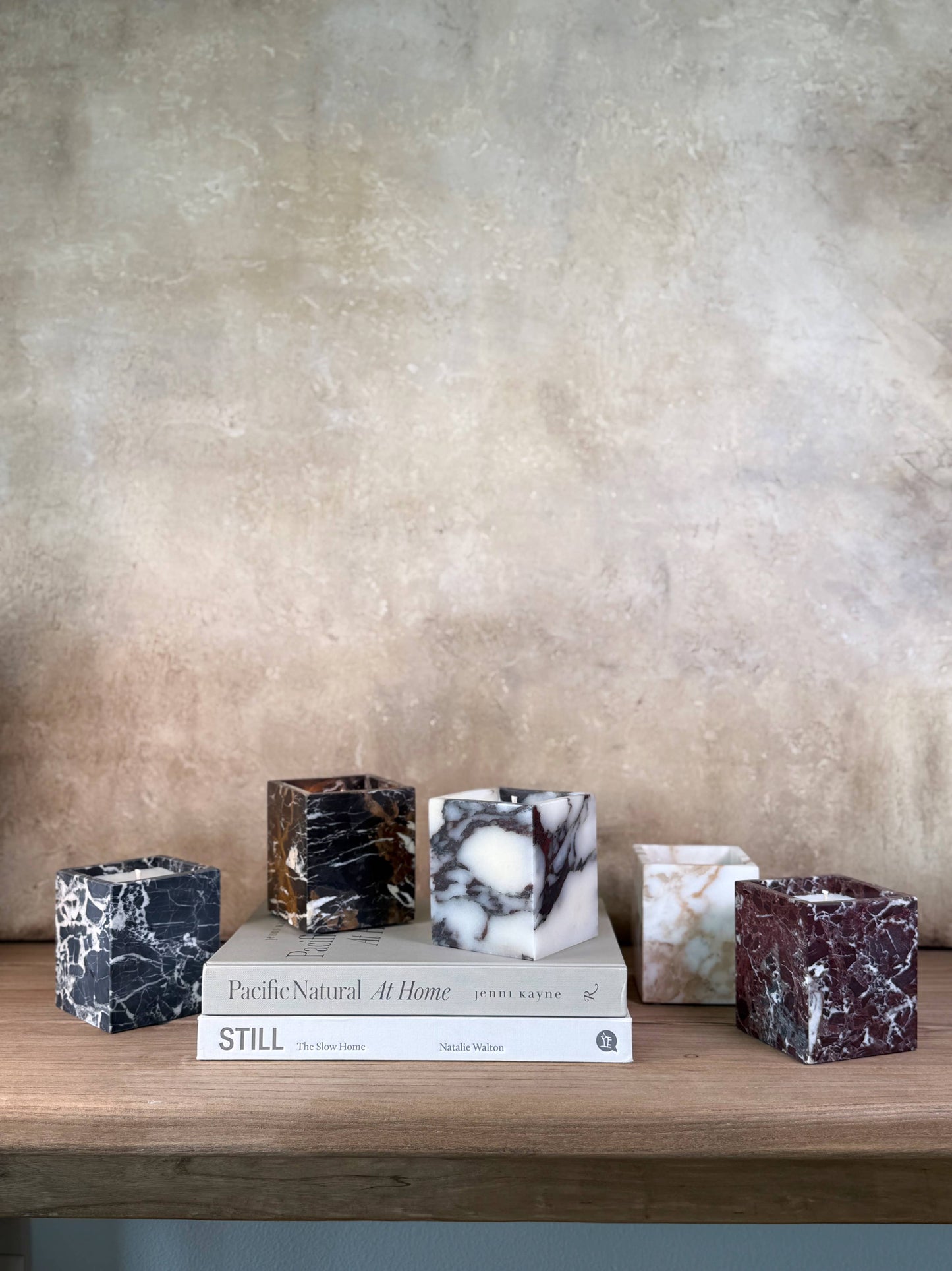 Five Marble Candles in assorted colors are displayed on a wooden shelf above two stacked books, Pacific Natural At Home and STILL, with a textured beige background.