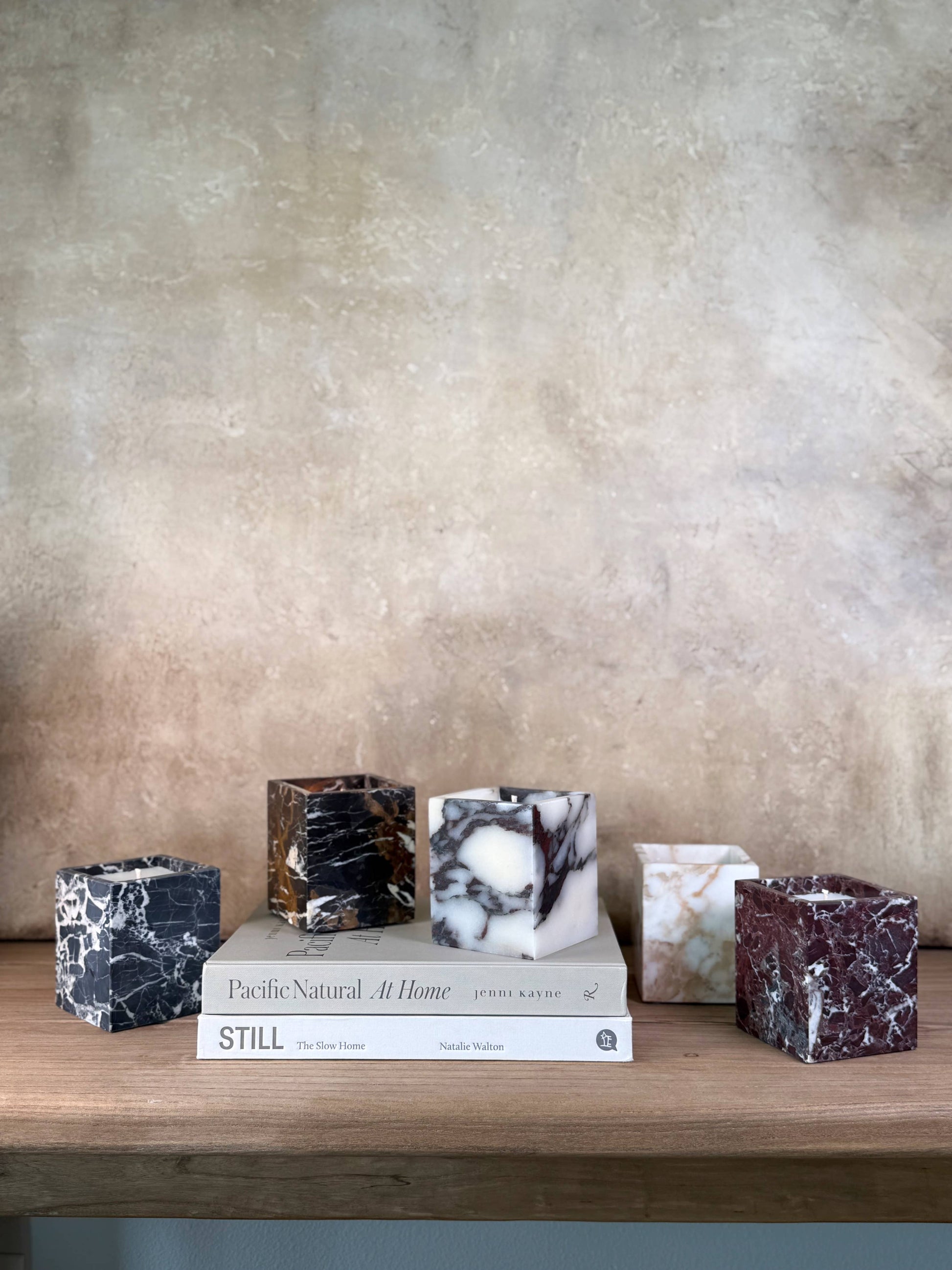 Five Marble Candles in assorted colors are displayed on a wooden shelf above two stacked books, Pacific Natural At Home and STILL, with a textured beige background.