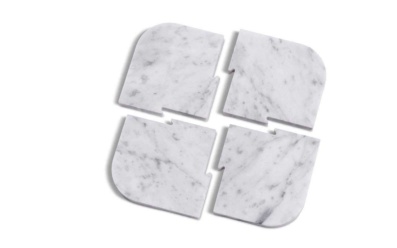 The Marble Coasters + Trivet set features four white marble coasters with rounded edges and side notches, arranged with small gaps to form a square—an elegant set ideal for display on a walnut stand.