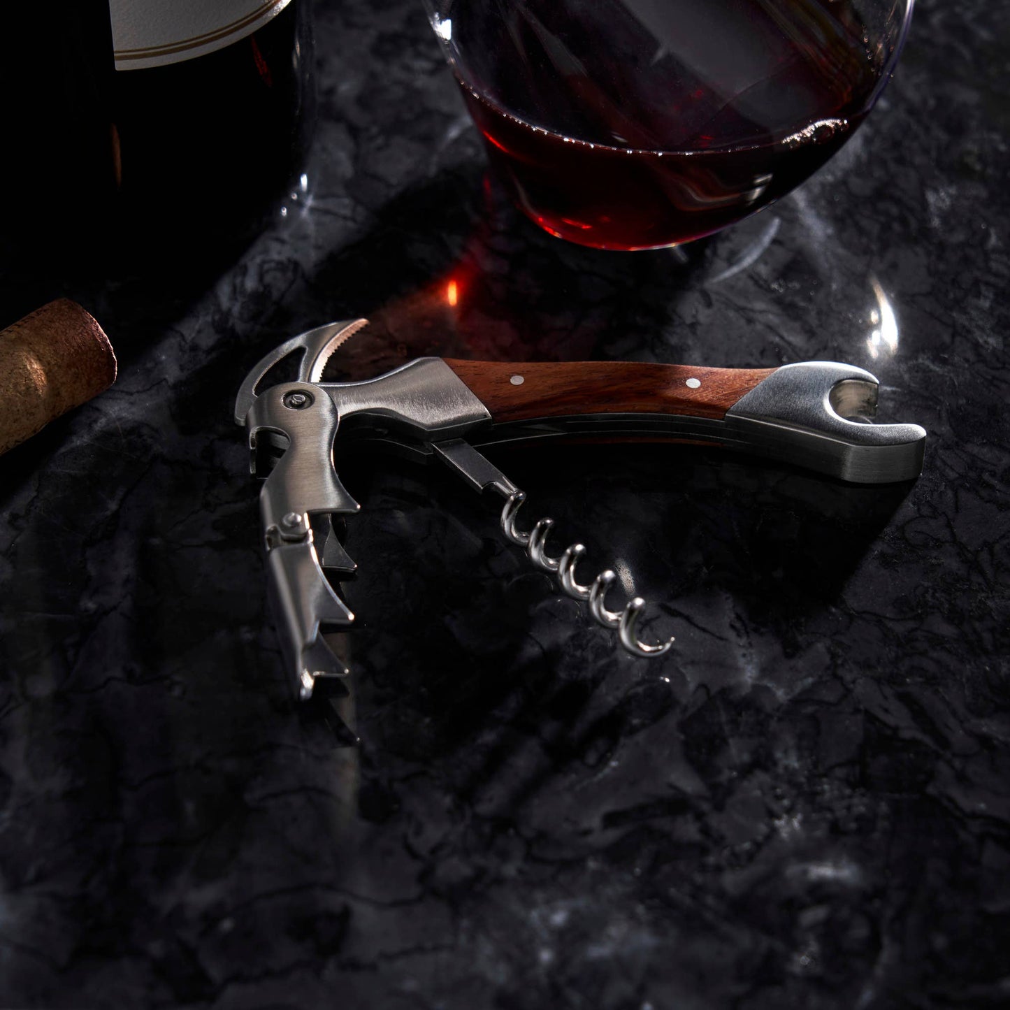 The Rosewood DoubleHinged Corkscrew and Foil Cutter rests on dark marble beside a glass of red wine, a wine bottle, and a cork.