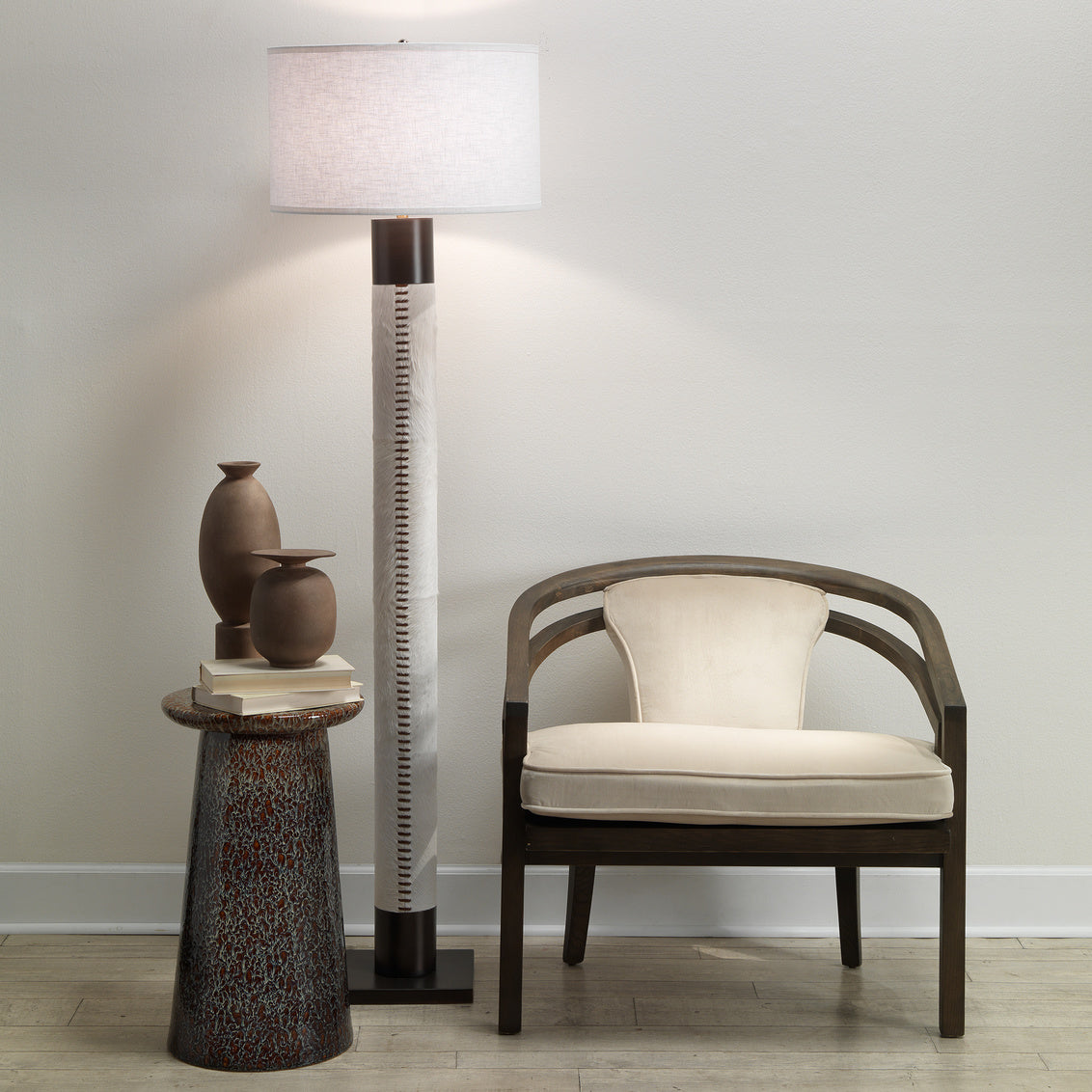 A modern room featuring the Stitched Cowhide Floor Lamp in rubbed bronze, a small round table with stacked books and two vases, and a wooden chair with a cream cushion, all set against a light-colored wall and wood floor.