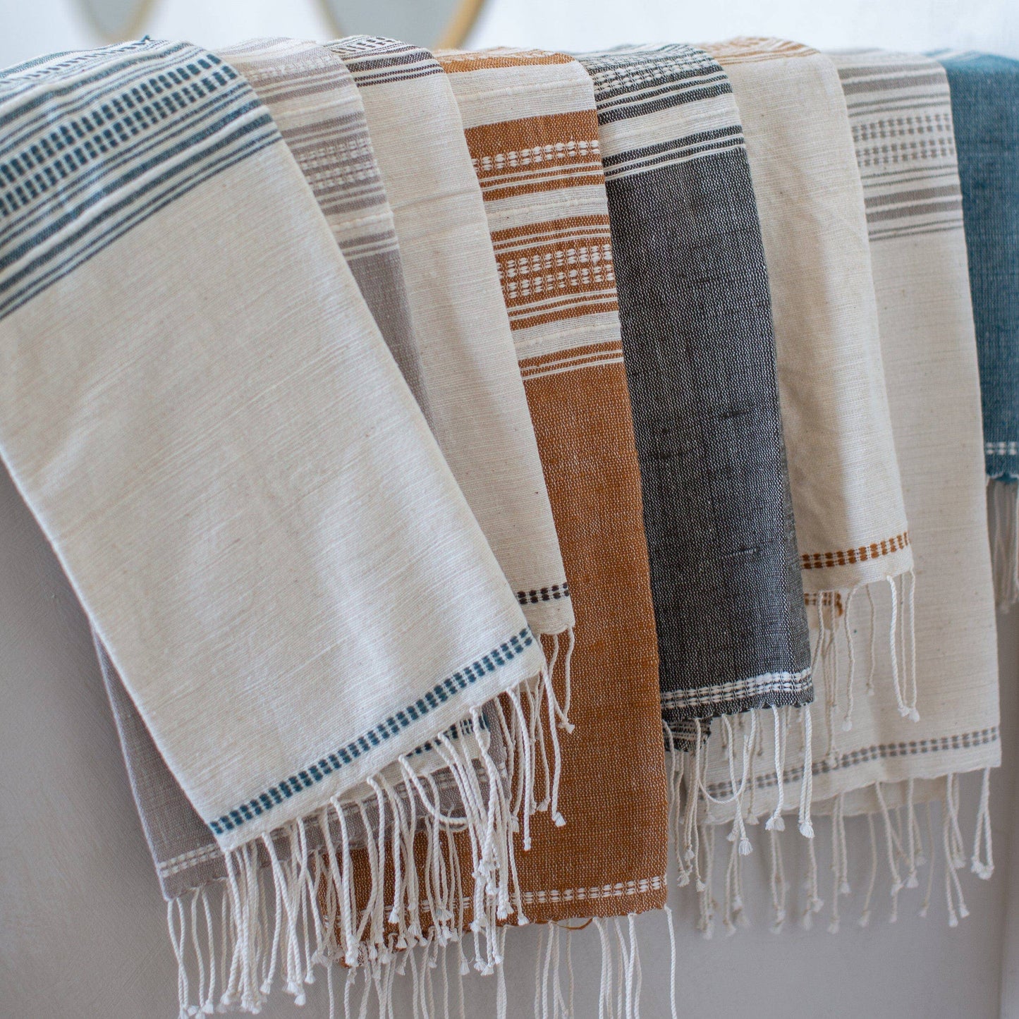 Five Handwoven Ethiopian Cotton Hand Towels with fringe hang side by side in beige, brown, gray, white, and blue striped designs. These eco-friendly towels bring artisanal charm to any space.