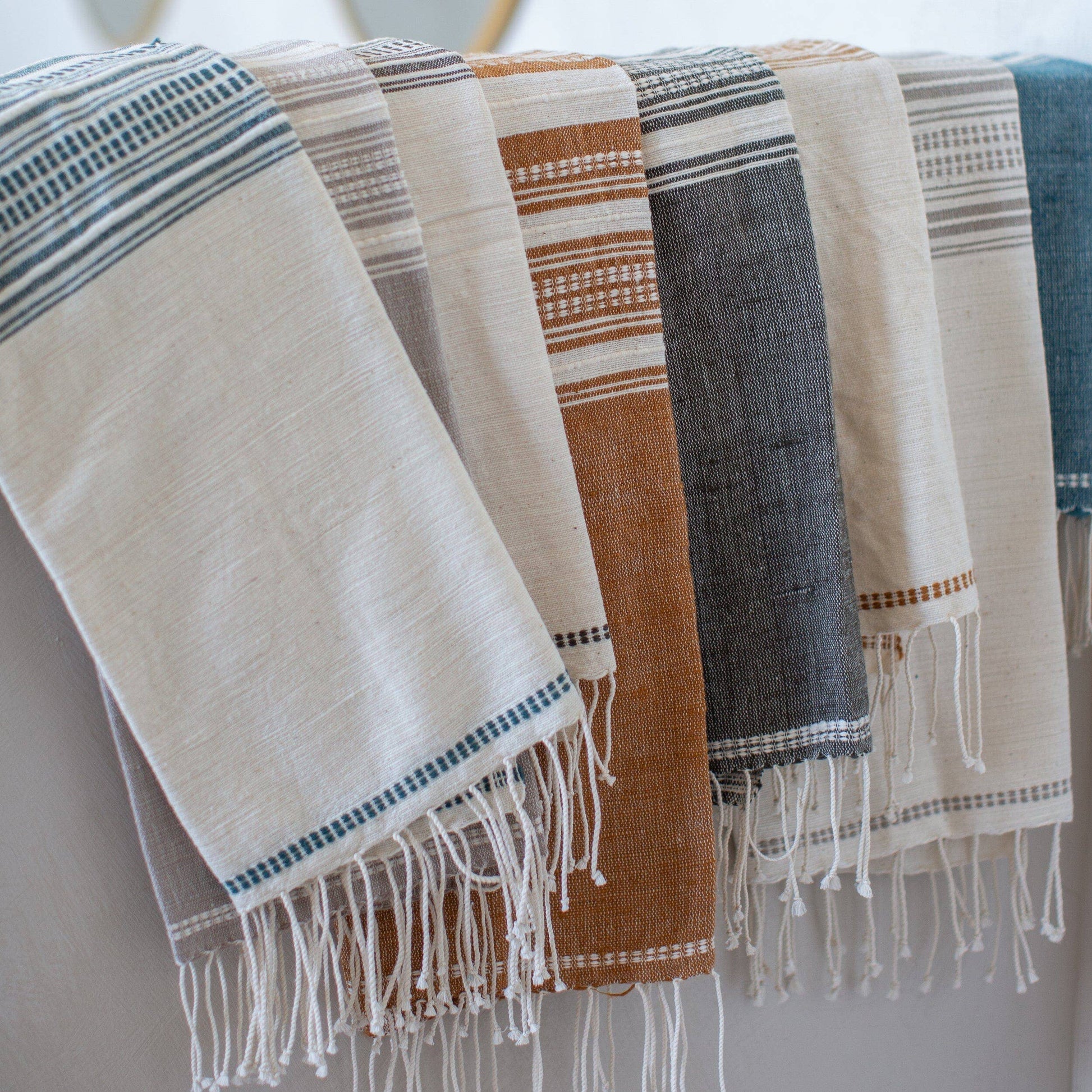 Five Handwoven Ethiopian Cotton Hand Towels with fringe hang side by side in beige, brown, gray, white, and blue striped designs. These eco-friendly towels bring artisanal charm to any space.