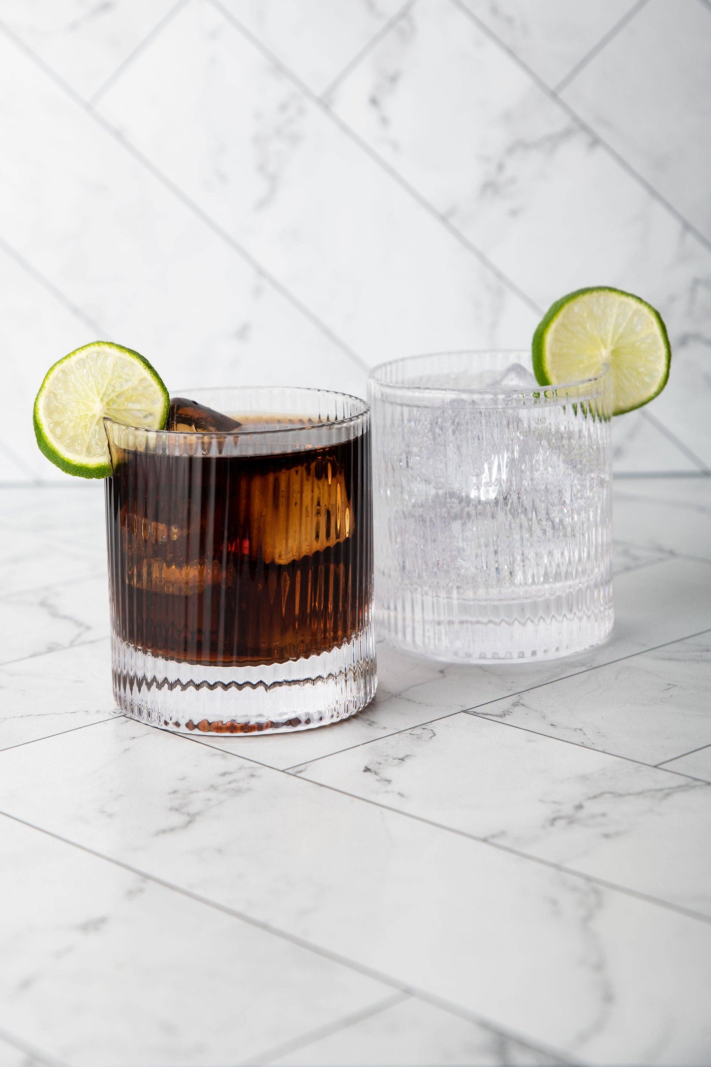 Two Vintage Straight Line Tumbler 10 oz glasses sit on white marble; one holds a dark drink with ice, the other a clear beverage. Both lead-free crystal tumblers are garnished with lime slices.