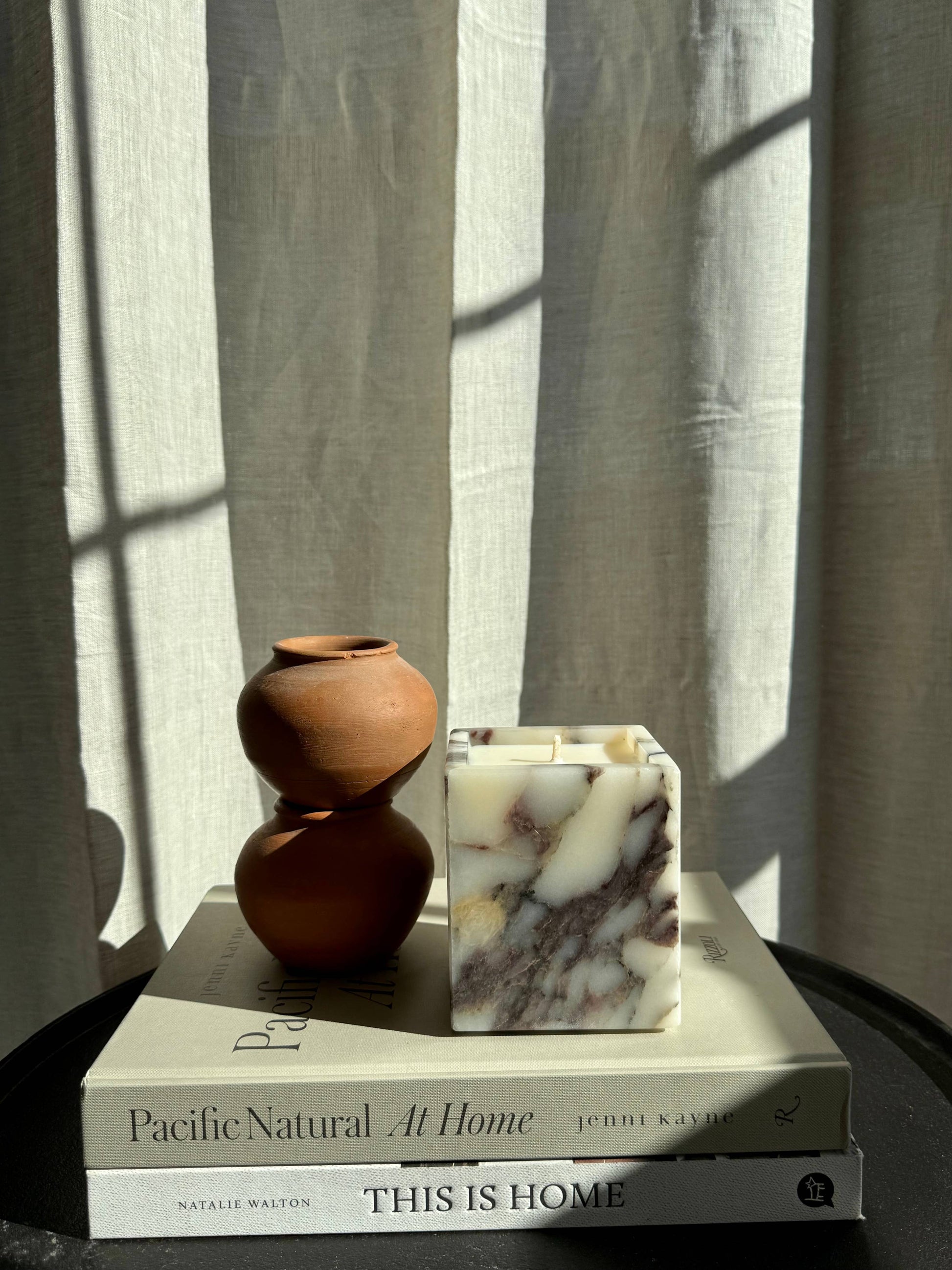 Two stacked small brown clay pots and a Marble Candle rest on two books—Pacific Natural: At Home and This Is Home—while sunlight casts soft shadows across a light fabric backdrop.