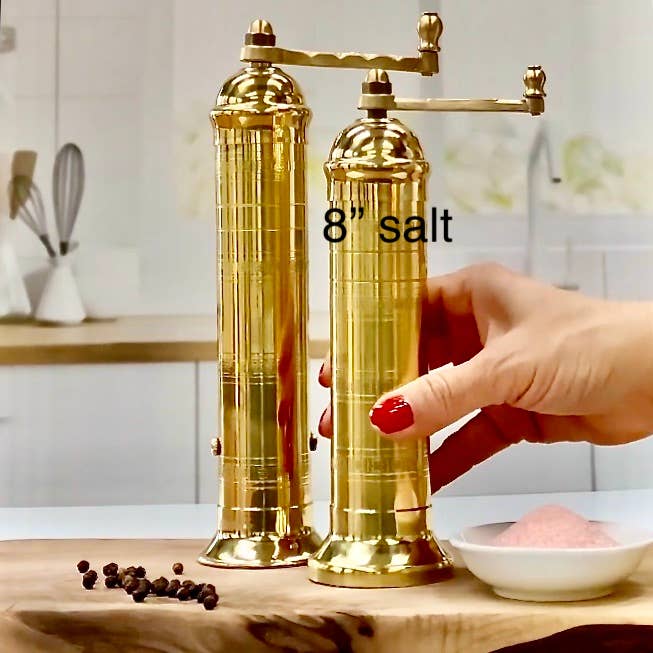 Two tall, shiny gold 'Brass Mill' 8" salt grinders are on a kitchen counter. A hand holds one labeled salt near a small dish of pink salt and scattered black peppercorns. The kitchen background is blurred.
