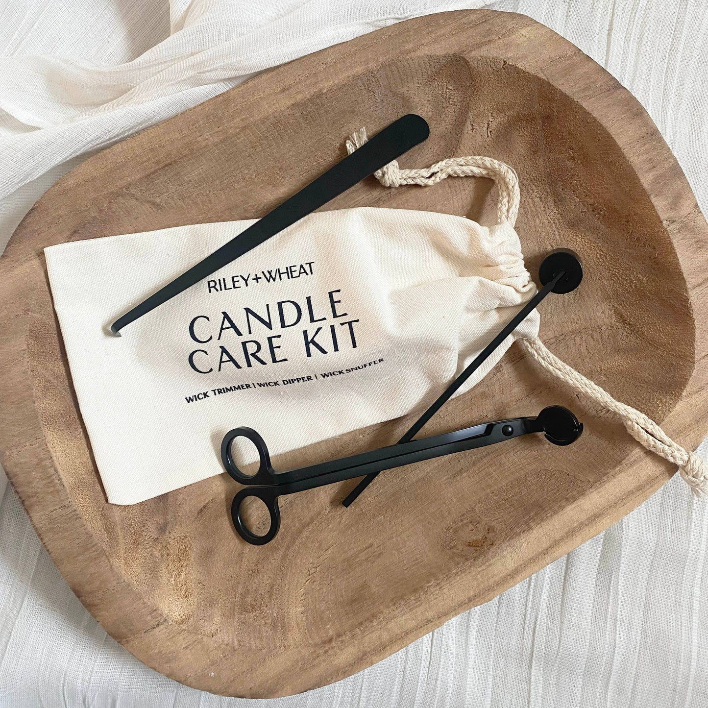 A wooden tray displays the Candle Care Kit, which includes a black metal wick trimmer, snuffer, dipper, and a drawstring bag, all neatly arranged on a light fabric surface.