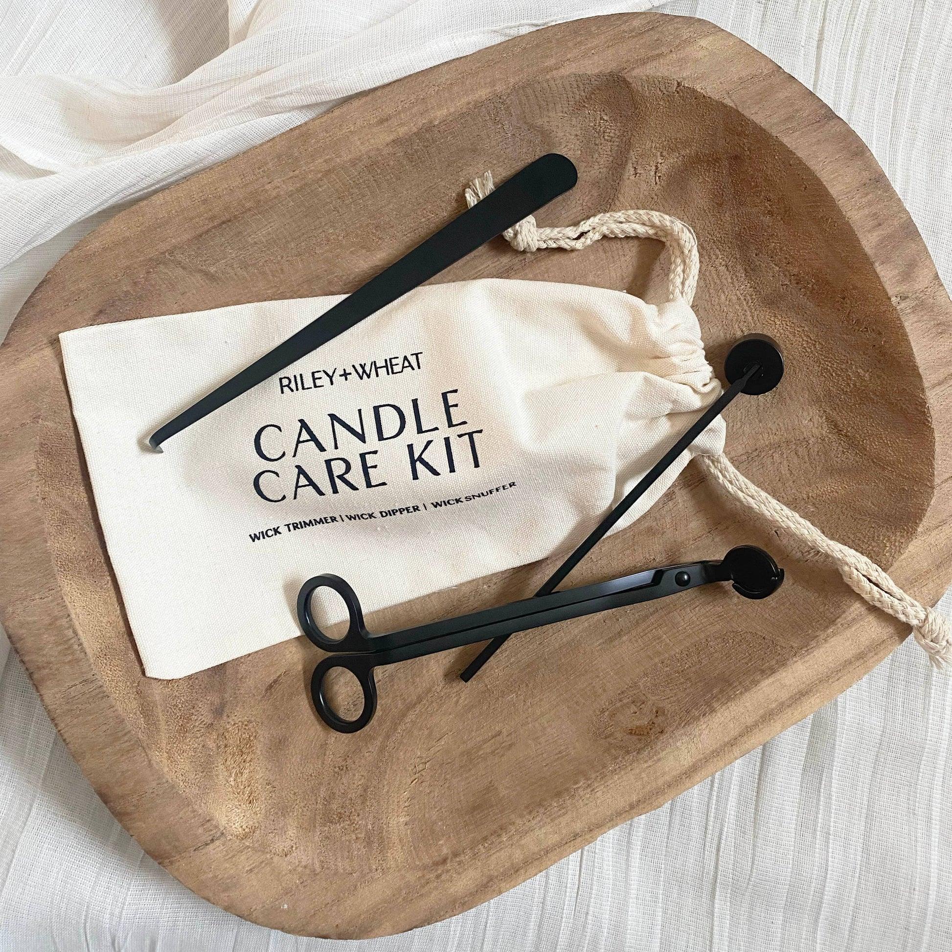 A wooden tray displays the Candle Care Kit, which includes a black metal wick trimmer, snuffer, dipper, and a drawstring bag, all neatly arranged on a light fabric surface.