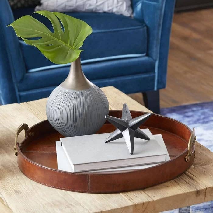 The Vintage Genuine Leather Tray holds a gray vase with a large green leaf, two stacked white books, and a black metal star sculpture on a coffee table in front of a blue upholstered chair, adding style to your living space.