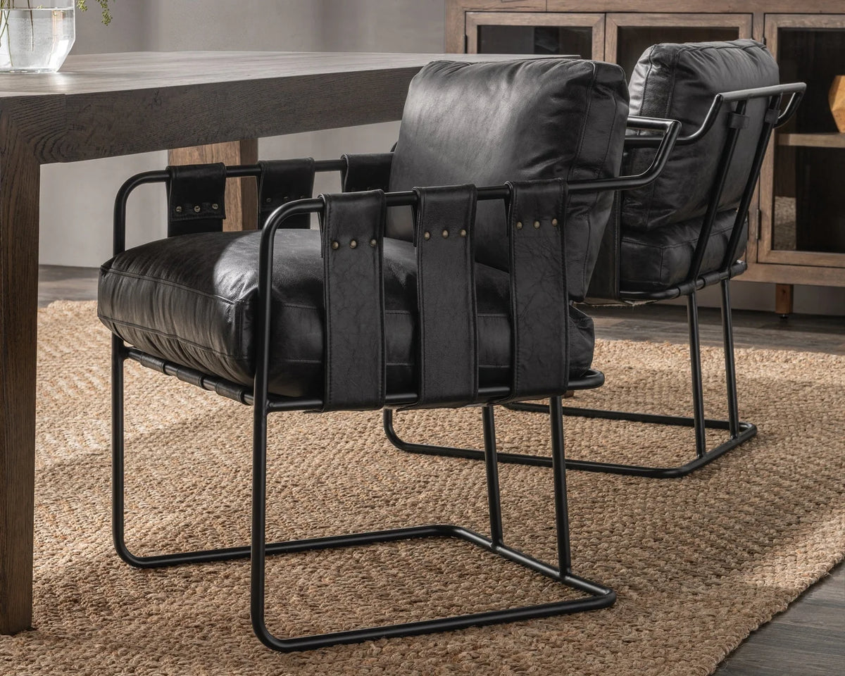 Two Strap Dining Chair (21.5″W x 26.5″D x 32″H) with metal frames sit beside a dark wooden table on a woven jute rug, with a cabinet and glass vase in the background.