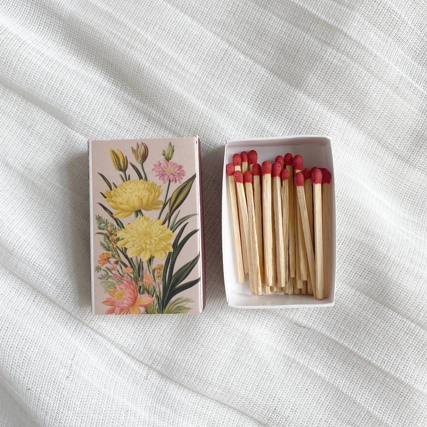 A Vintage Matchbook with a floral design sits open on textured white fabric, displaying red-tipped strike-on-box matches neatly arranged inside.
