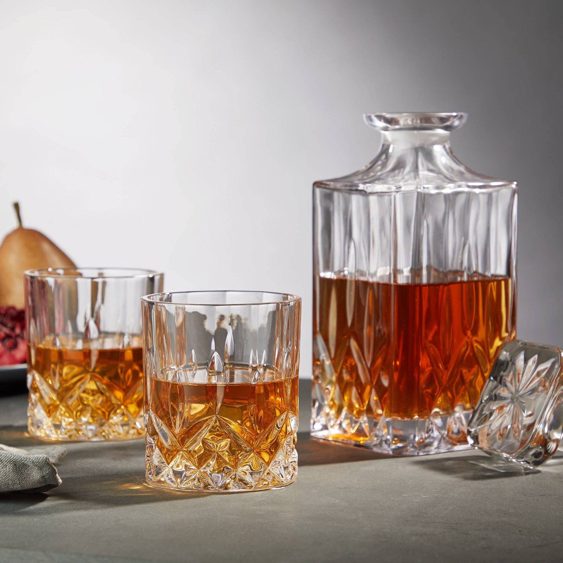 The Crystal Whiskey Decanter and Tumblers (Set of 3) are shown on a grey surface, filled with whiskey, accompanied by a pear and pomegranate in the background. The decanter's stopper rests beside the matching glasses.
