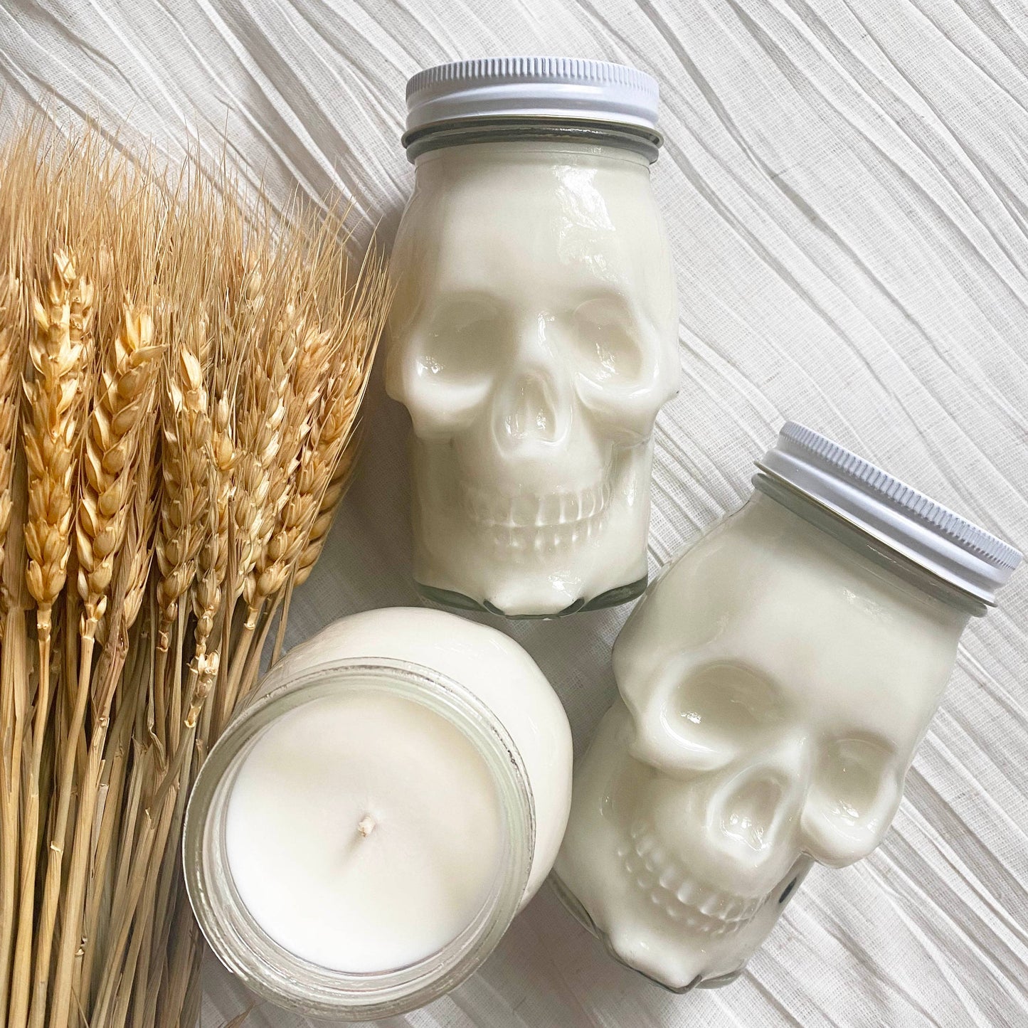 Two white Skull Candles with white lids and a round soy wax jar candle are displayed beside wheat stalks on textured white fabric, creating a cozy fall-inspired decor setup.
