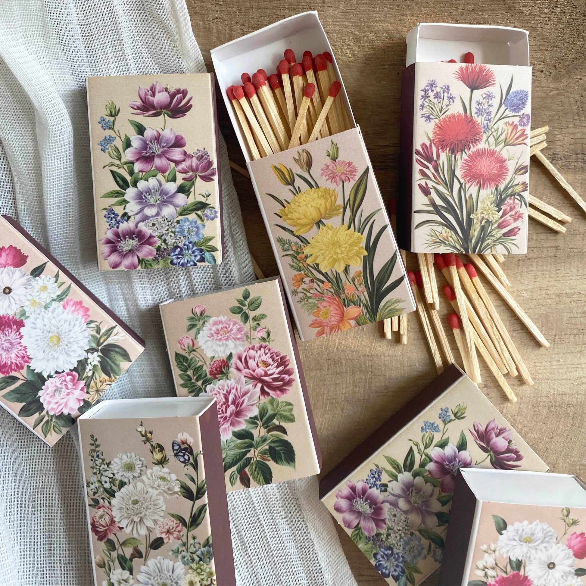 Vintage Matchbooks with colorful floral designs are arranged on a wooden surface, some open to display red-tipped strike-on-box matches, while others remain closed. A white fabric is partially visible beneath them.