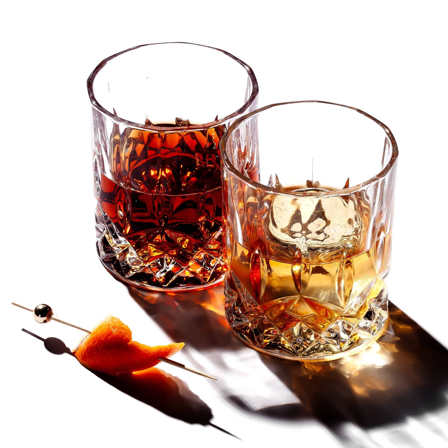 A Crystal Cut Old Fashioned Whiskey Glass 10oz sits on a white surface with drinks inside, next to a cocktail pick garnished with orange peel. Its beautiful reflections make it an ideal gift for whiskey lovers.